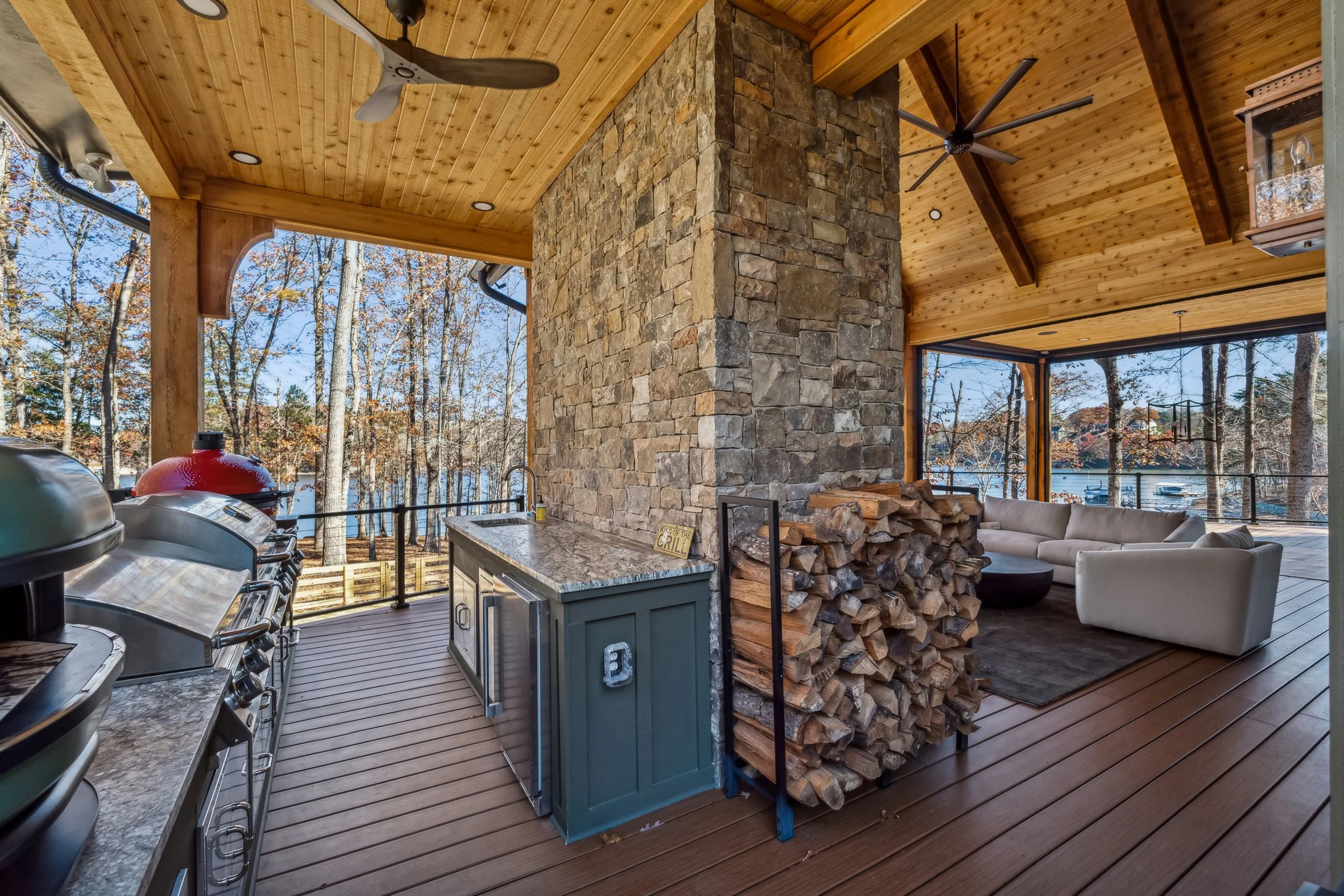 Covered outdoor living space with a stone fireplace, wood ceiling, ceiling fans, a sofa, and a view of trees and a lake outside.