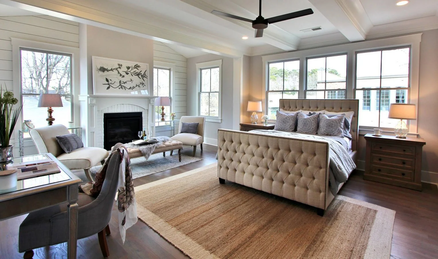 A spacious living room with a large bed, tufted headboard, multiple windows, a fireplace, tasteful lamps, and a dining area with a desk and chairs, all decorated in neutral tones with elegant furnishings.