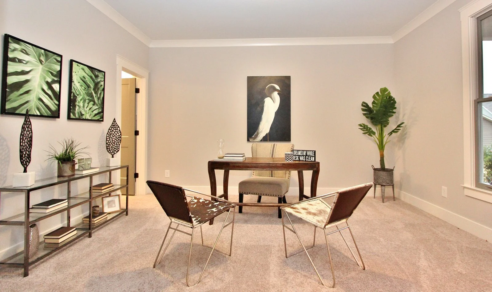 Living room with beige carpet, a desk and chair, wall art of a bird, a large plant in a pot, and decor items including books and art pieces.