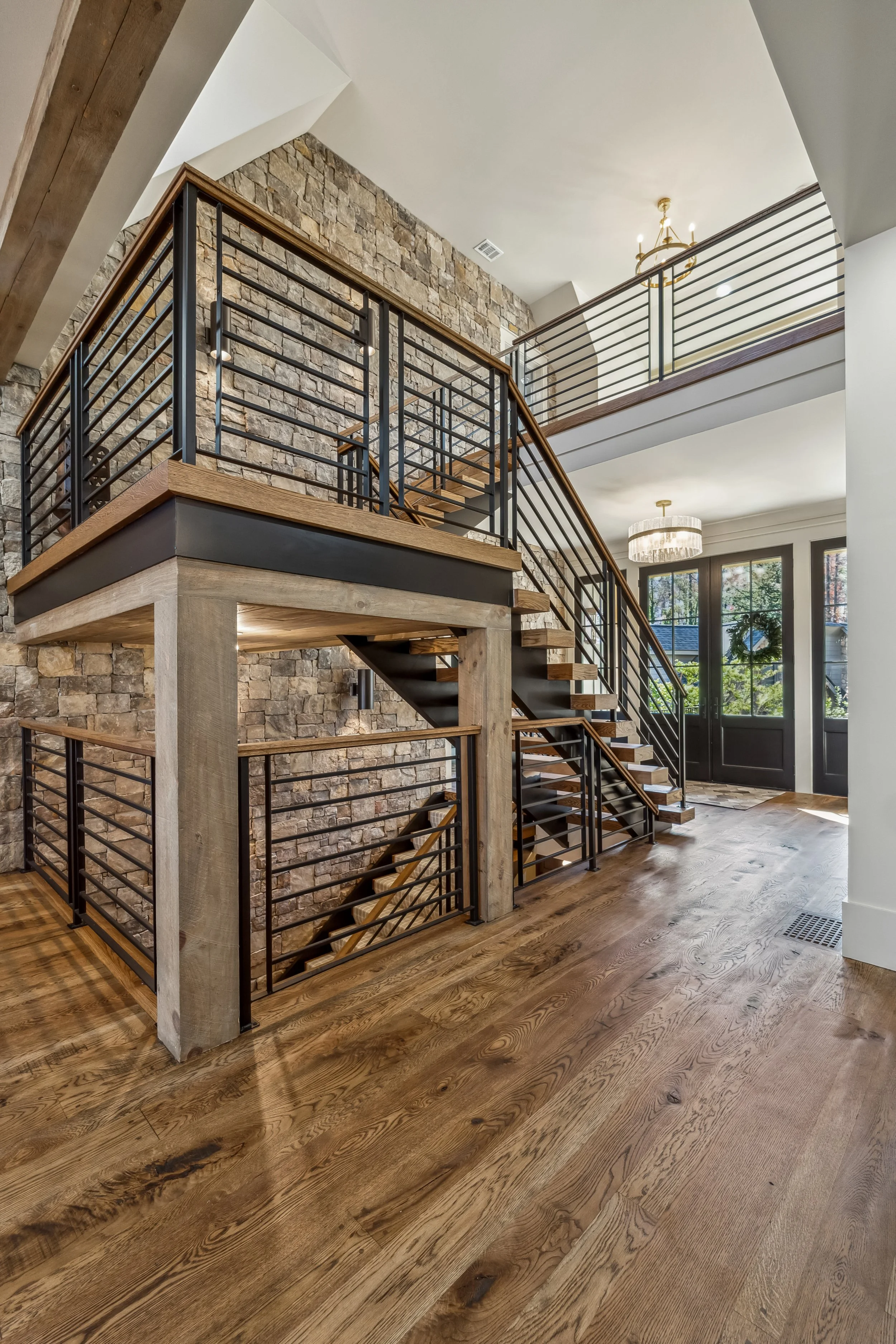 Interior view of a modern house with a wooden staircase and black metal railings. The wall behind the staircase is made of stone, and there are large windows in the background letting in sunlight. The ceiling has a chandelier, and the floor is wooden