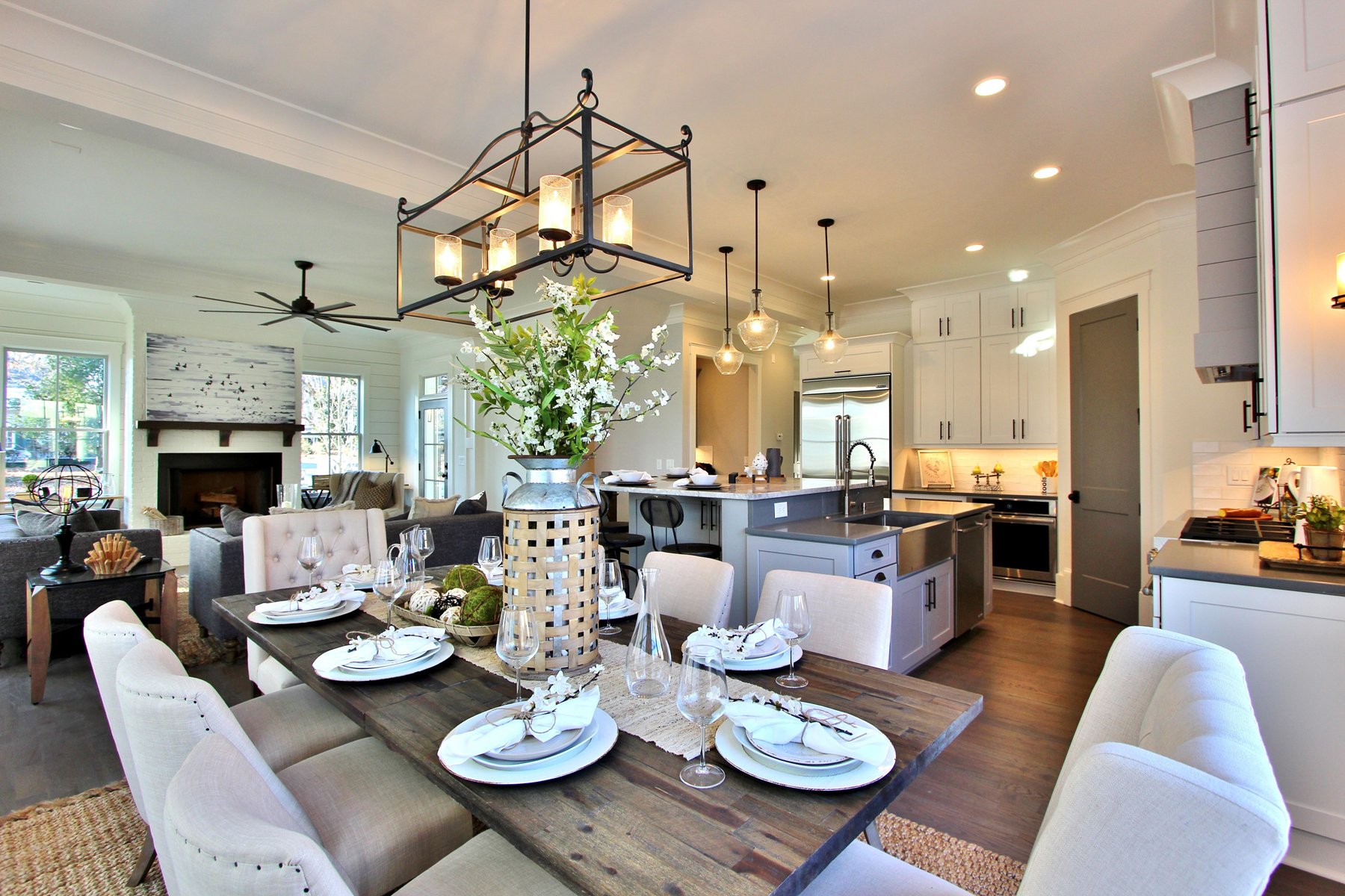 Open-concept kitchen and dining room with white cabinetry, stainless steel appliances, a large wooden dining table set for a meal, and a living area with a fireplace and comfortable chairs.