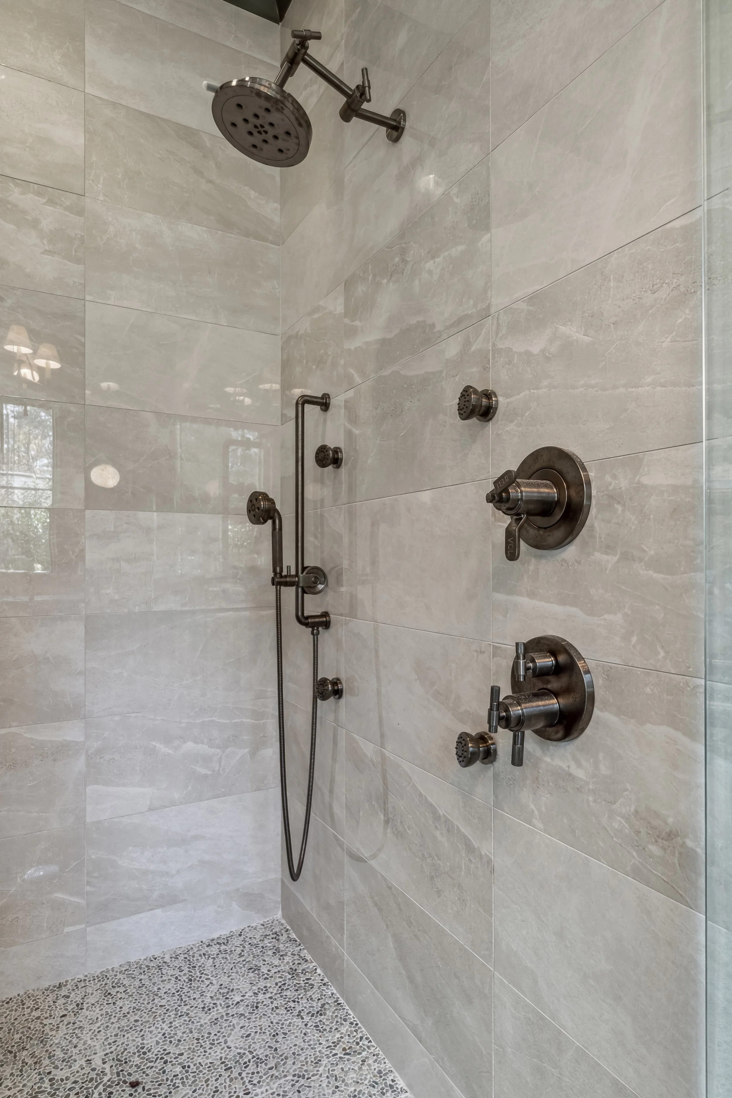Indoor shower with multiple showerheads and controls, tiled walls and pebble-textured floor.