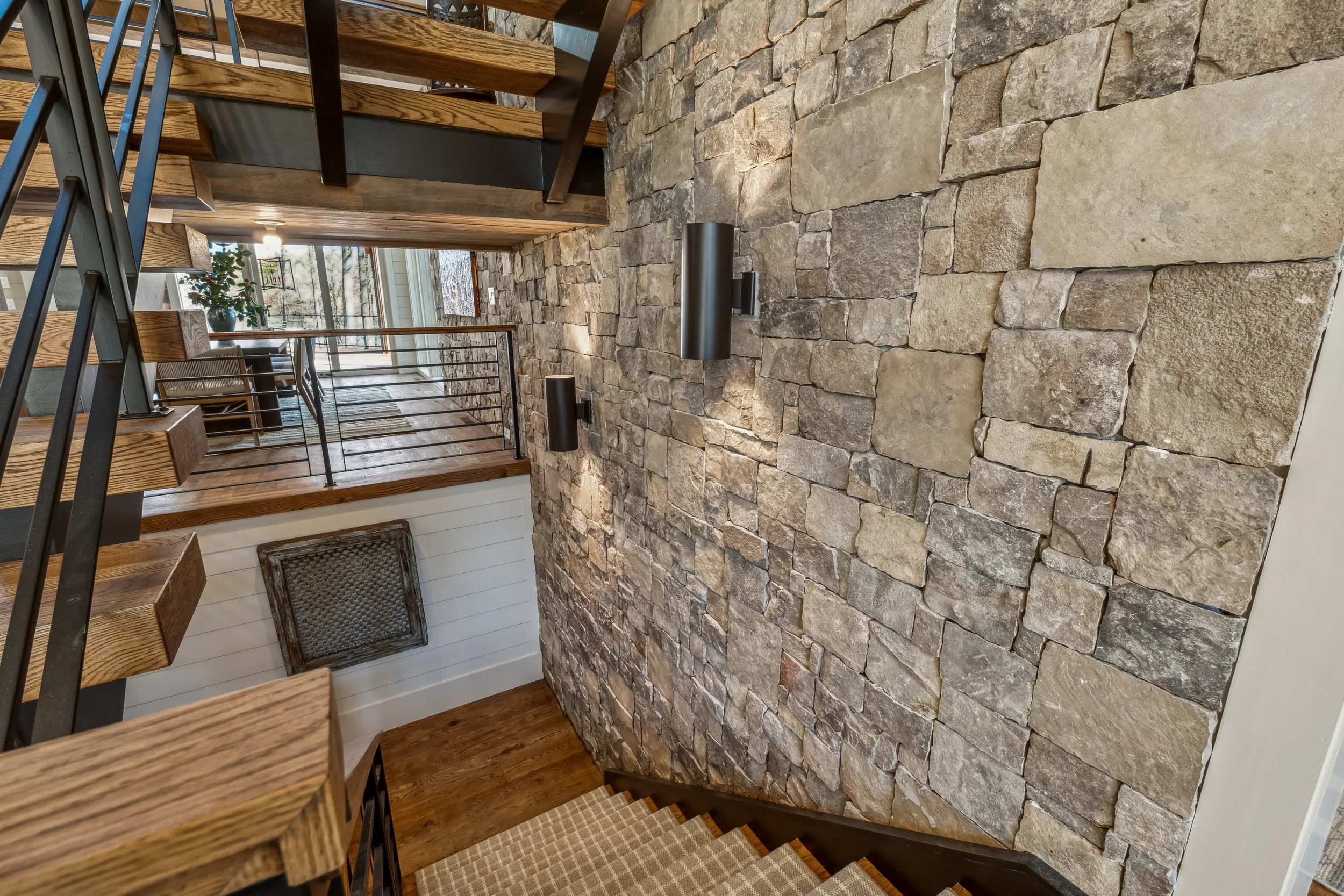 Interior view of a house showing a staircase with wooden steps and black metal railing, next to a stone accent wall, with a hallway leading to a sunlit room with large windows.