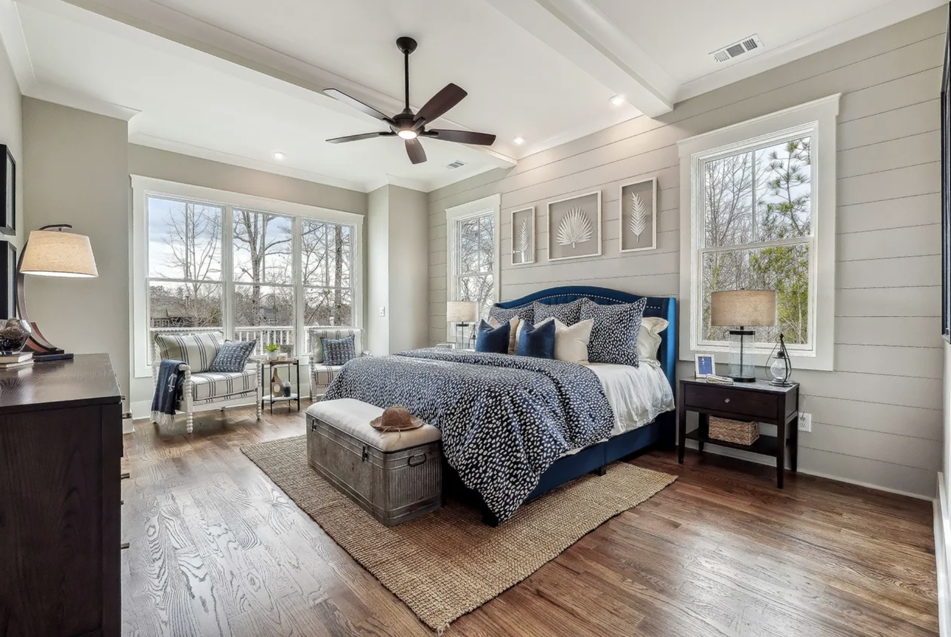 A spacious bedroom with large windows showing trees outside. The room features a blue bed with a navy and white patterned duvet and multiple pillows, a small dark bedside table with a lamp, framed botanical artwork on the wall, a bench at the foot of