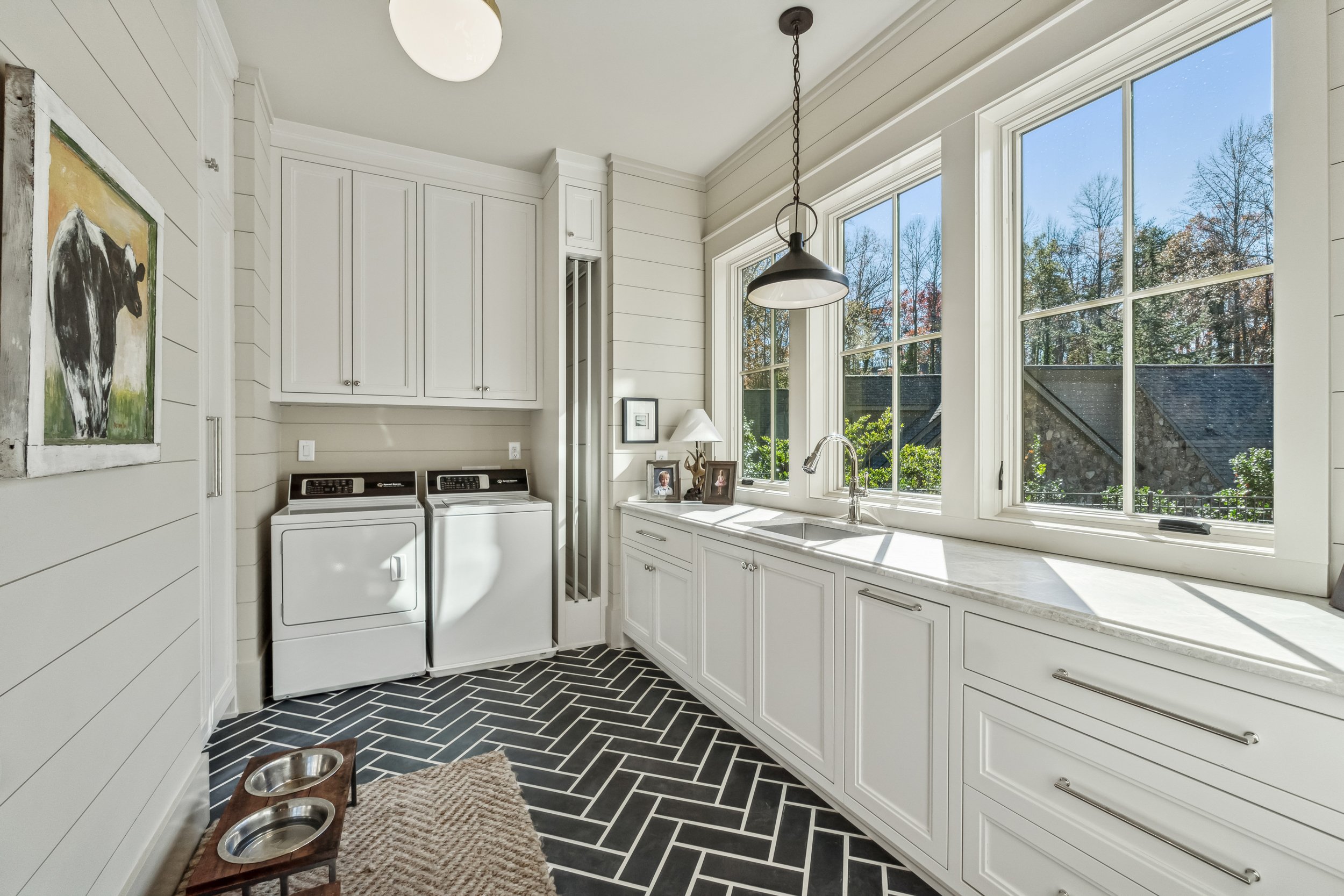 Bright laundry room with large windows, white cabinets, black and white herringbone tile floor, washing machine, and a small feeding station with two bowls.