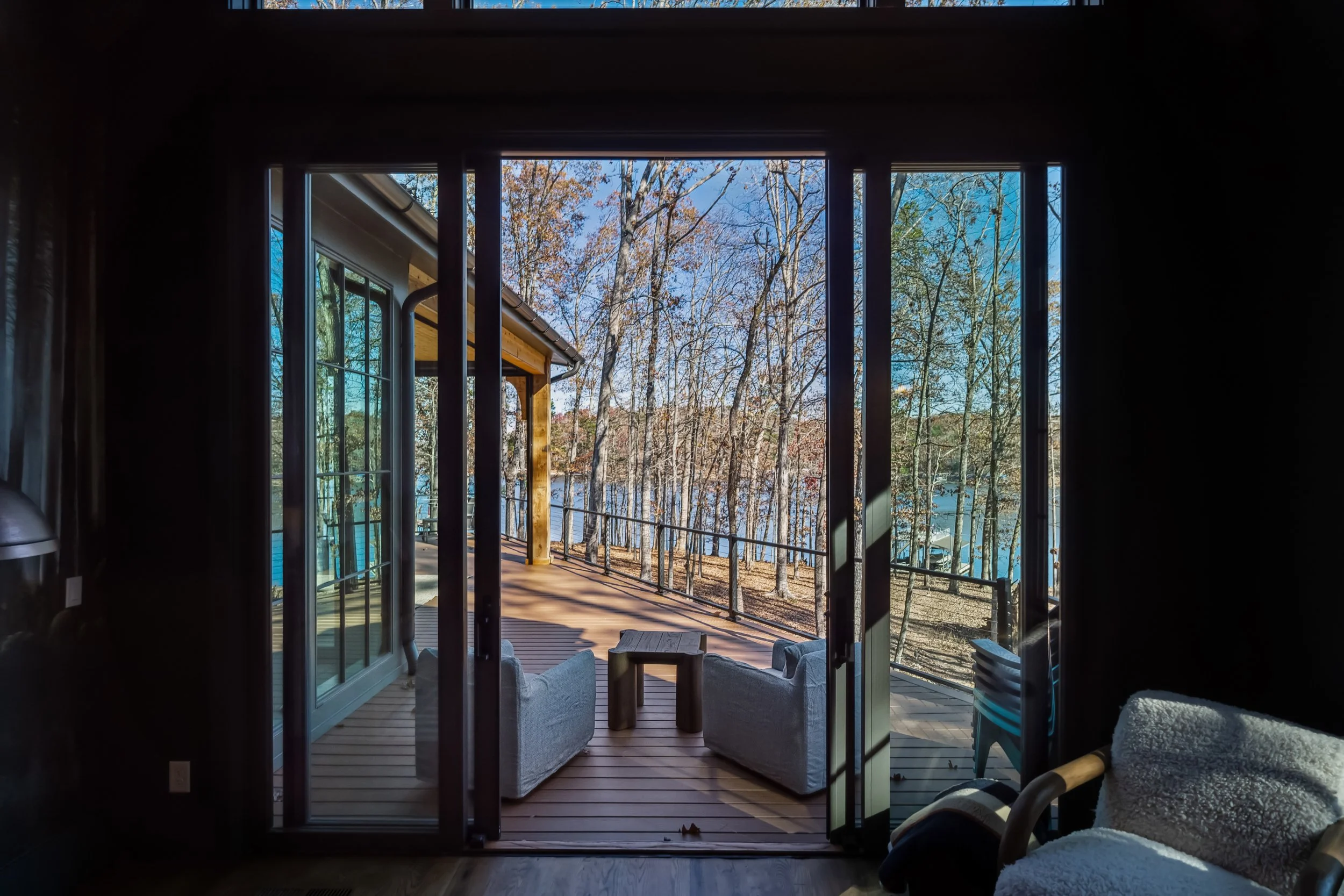 View from inside a house looking out through glass doors to a wooden deck and wooded area with leafless trees and a body of water in the background during fall.