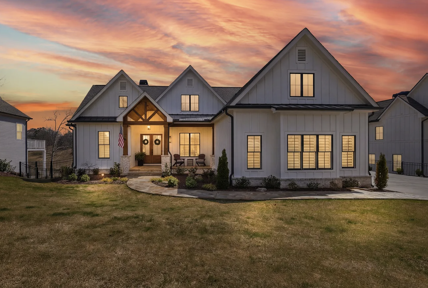 A modern white house with multiple gabled roofs and large windows, decorated with wreaths on the front door, is illuminated from within during sunset, with a landscaped yard and a paved pathway leading to the entrance.