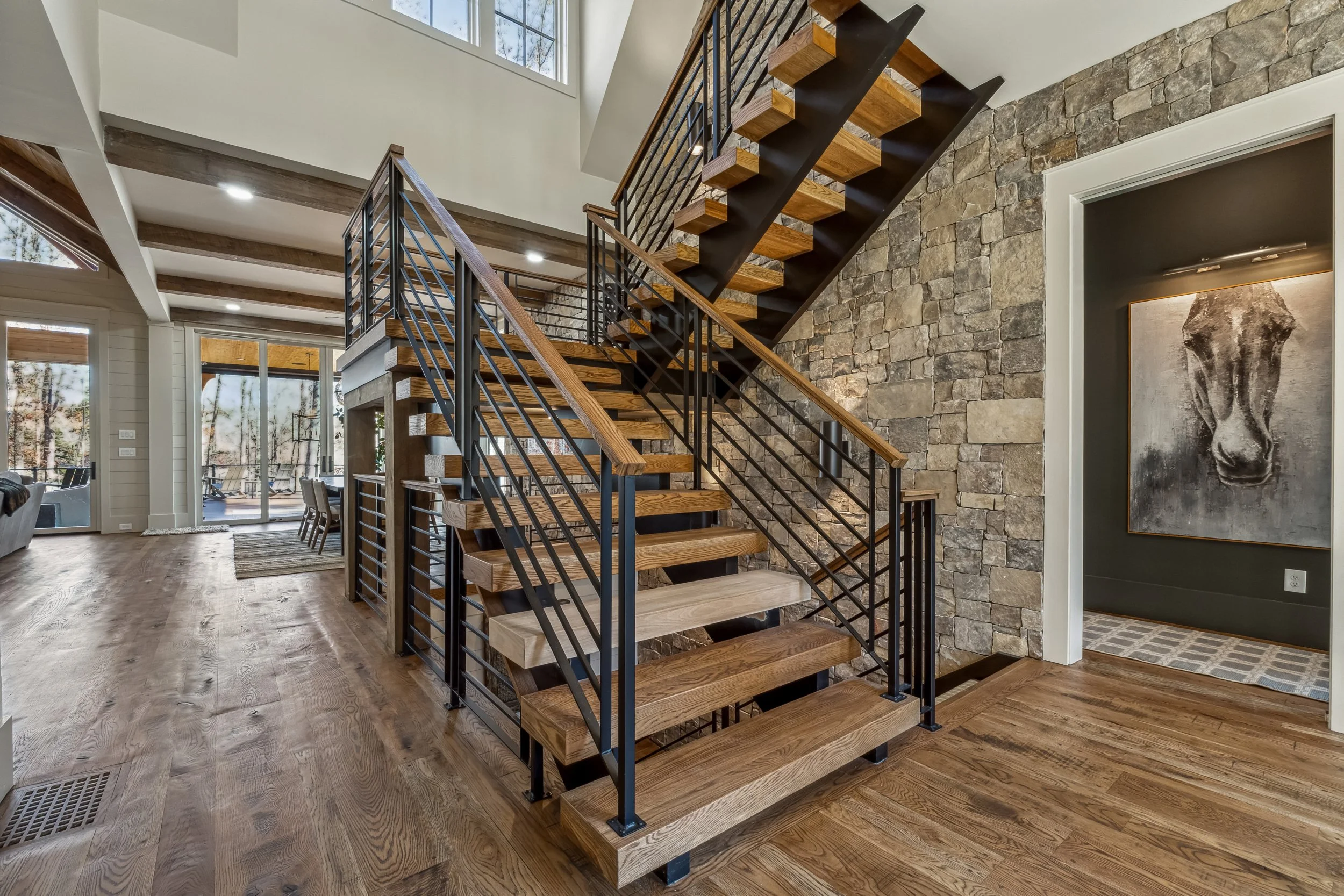Interior view of a modern home with a wooden staircase featuring black metal railings, a stone accent wall, large windows, and hardwood floors.