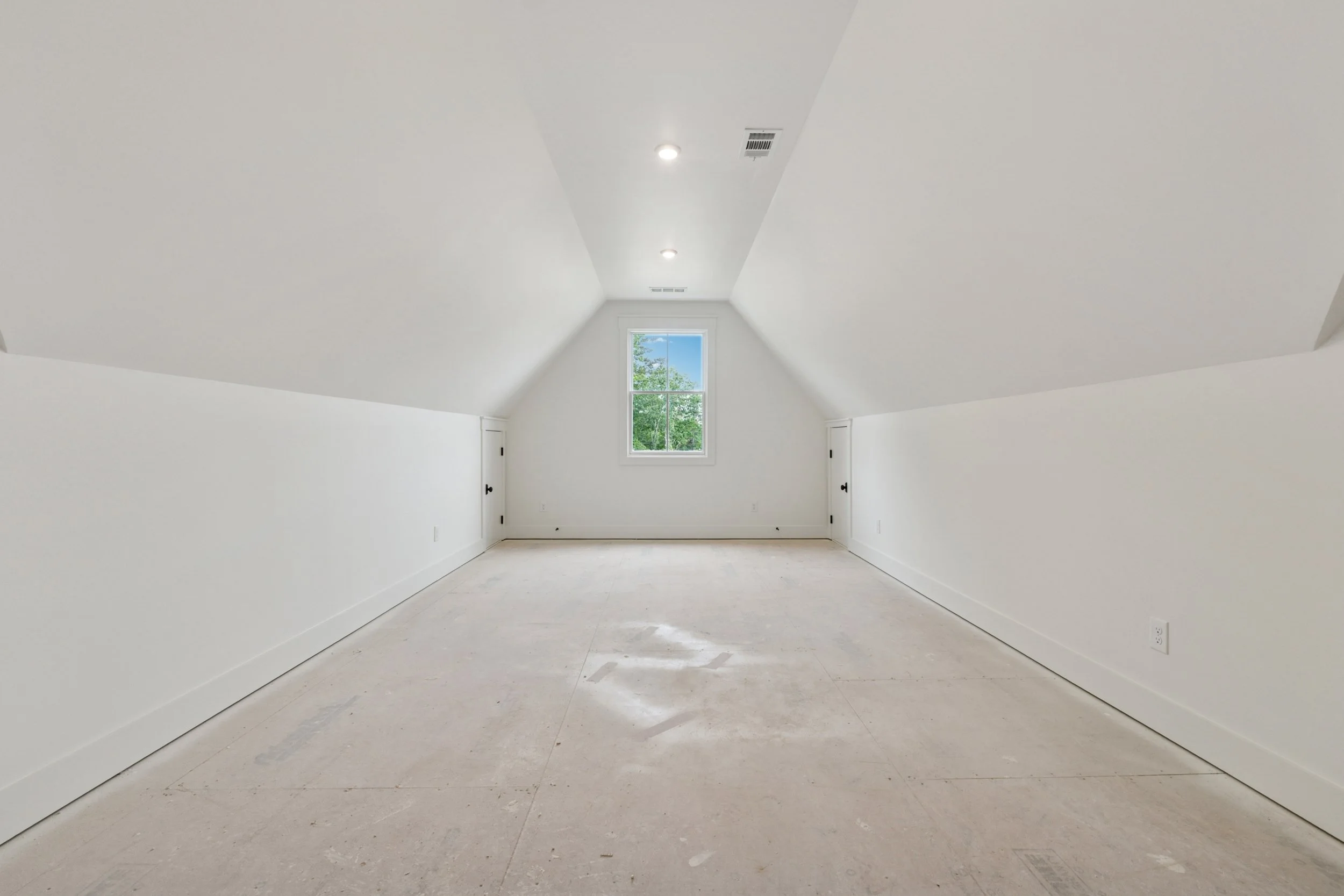 Empty attic room with white walls, ceiling, and trim, a window showing green trees outside, and two small doors on either side of the window.