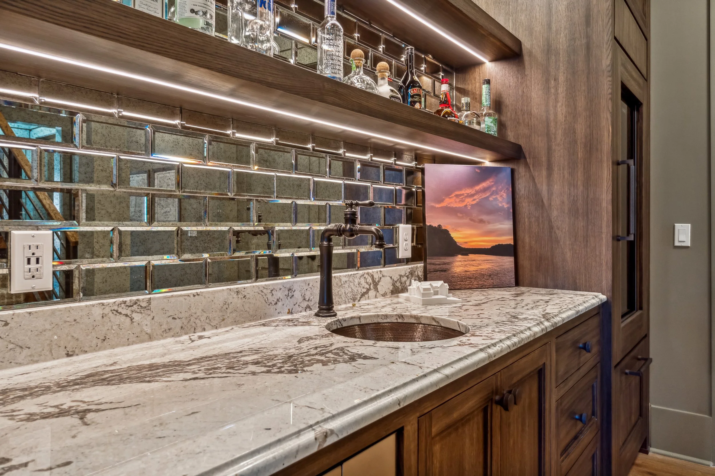 View of a kitchen counter with a marble surface, a black faucet, a miniature architectural model, and a wall-mounted television showing a sunset scene over water. Above the counter are wooden shelves with liquor bottles and decorative tiles.