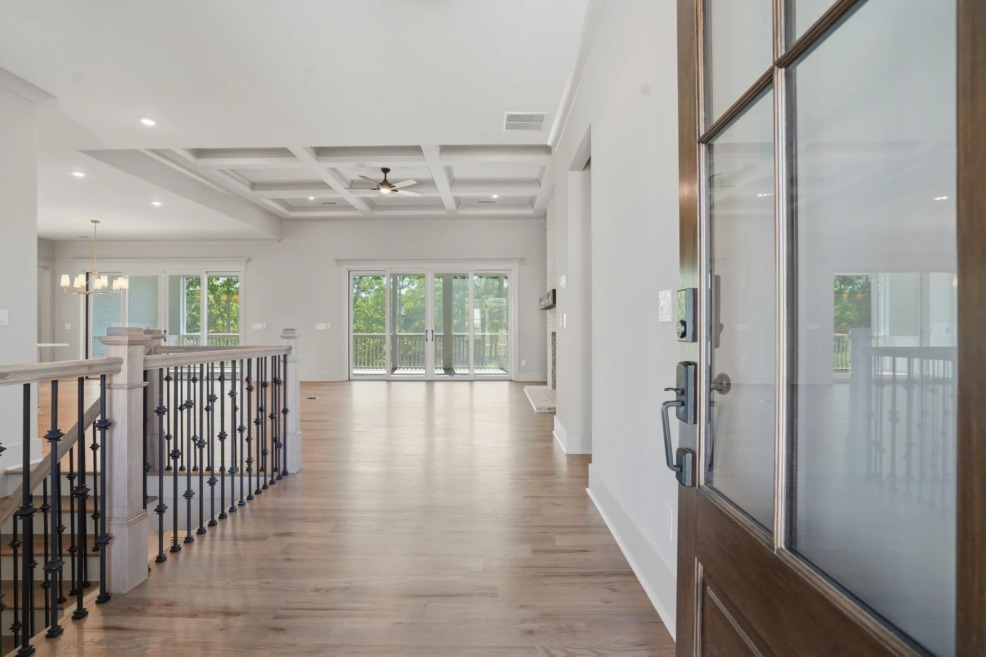 Open front door leading to a bright living room with wood flooring, large windows, a sliding glass door to a balcony, a ceiling fan, and a staircase with decorative black iron railing.