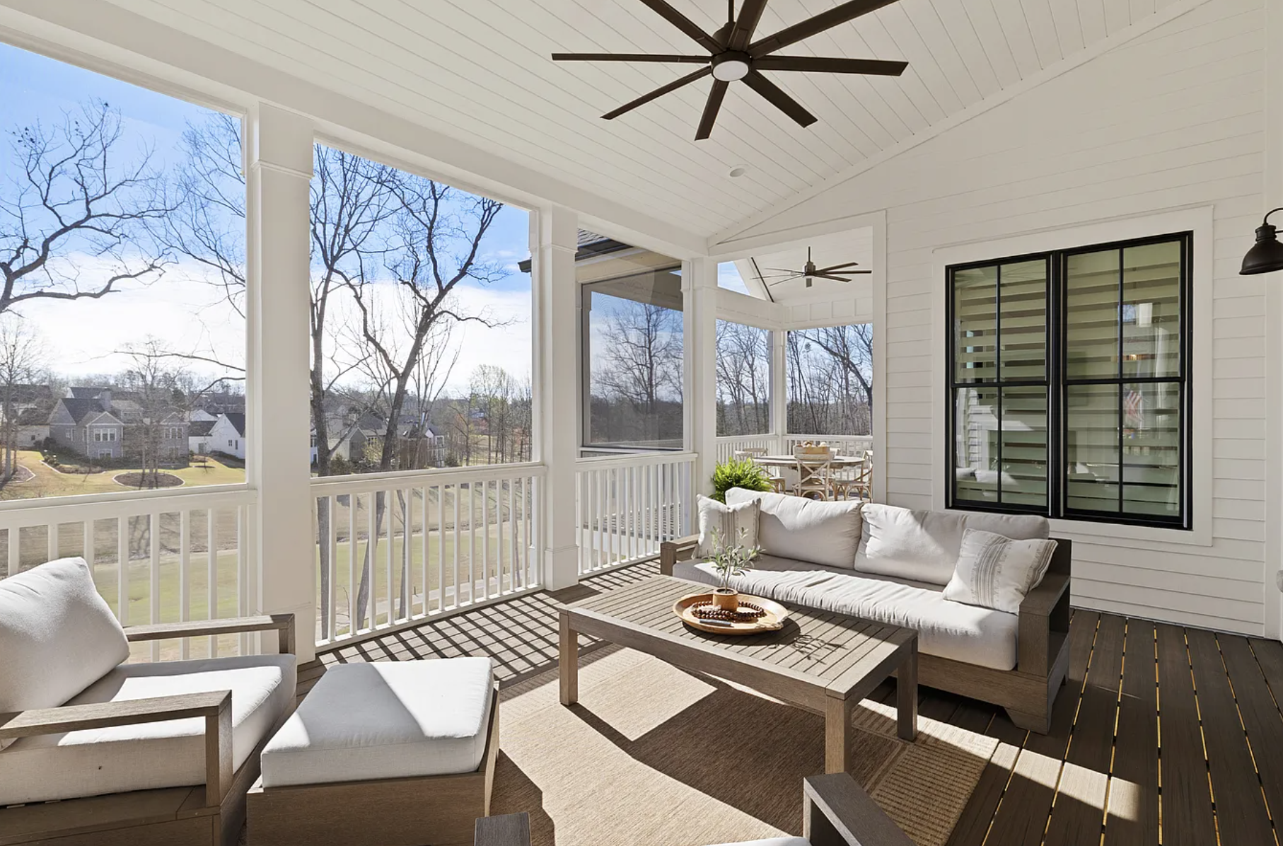 A porch with white railings and a ceiling fan, featuring outdoor seating including a cushioned sofa and chairs, overlooking a yard with leafless trees and neighboring houses under a clear blue sky.