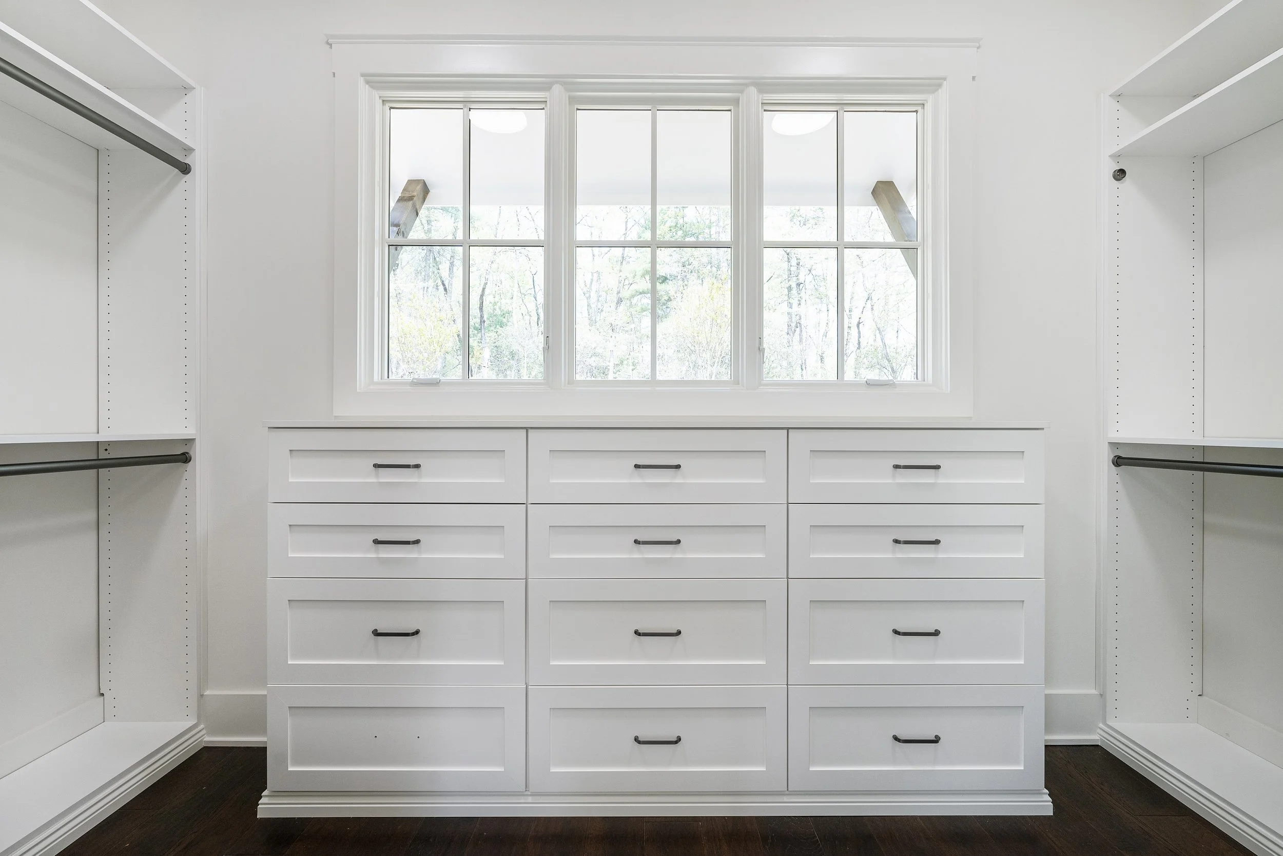 A spacious walk-in closet with white shelves and drawers, large window with views of trees, and dark wood floor.