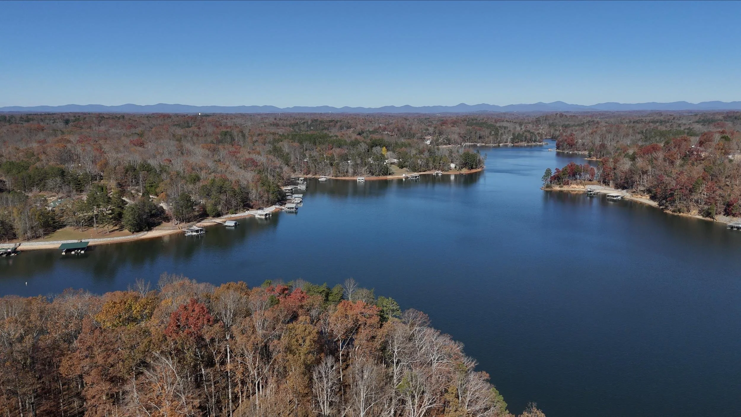 Aerial view of a large lake with a wooded shoreline in autumn, with boats docked along the edge and distant mountains on the horizon under a clear blue sky.