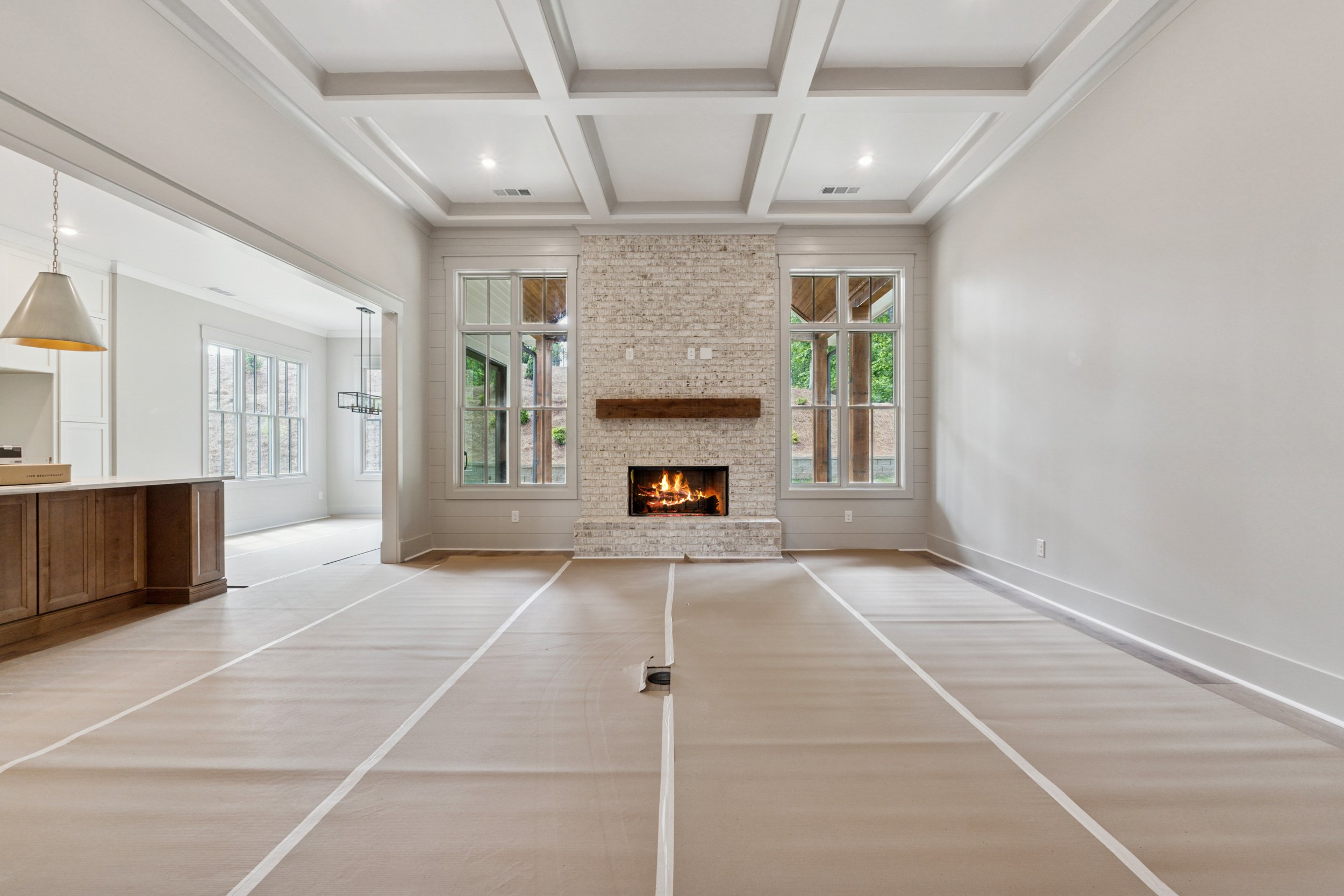 Empty living room with a white brick fireplace, large windows, and a coffered ceiling, with construction markings on the floor.
