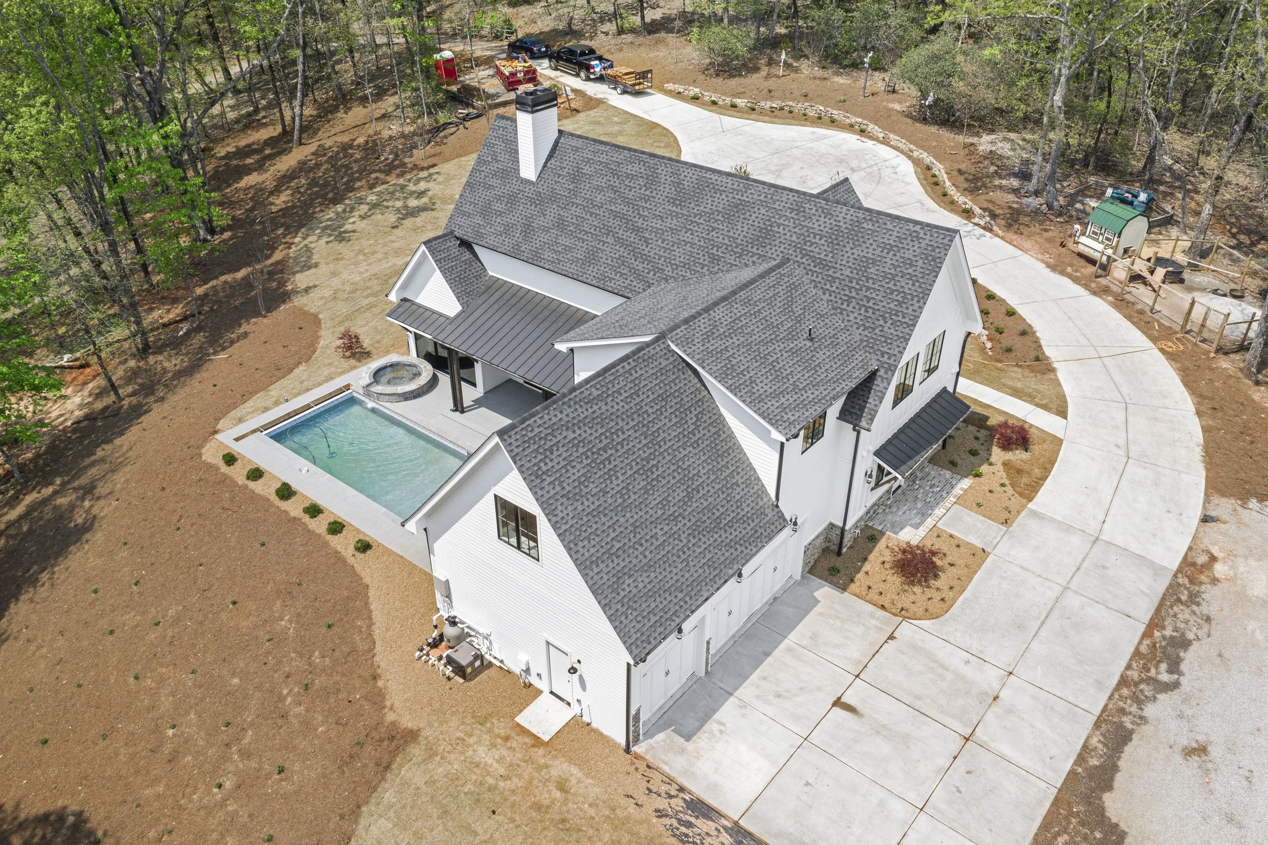 Aerial view of a modern white house with a gray shingled roof, an outdoor swimming pool, and a paved driveway surrounded by landscaped yard and trees.