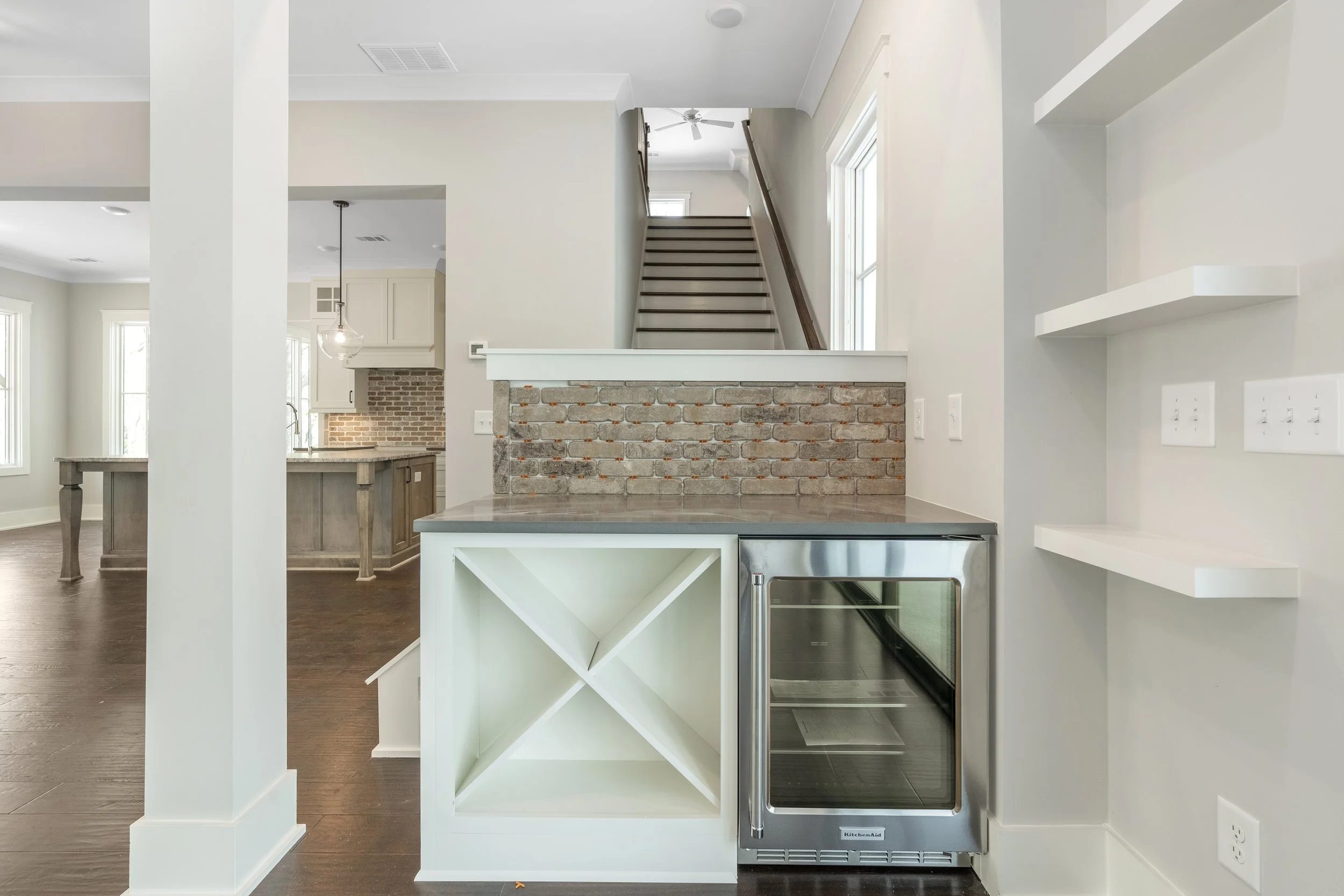 Interior view of a modern kitchen with white and beige cabinets, a brick partial wall, and a staircase leading upstairs. There are large windows letting in natural light.