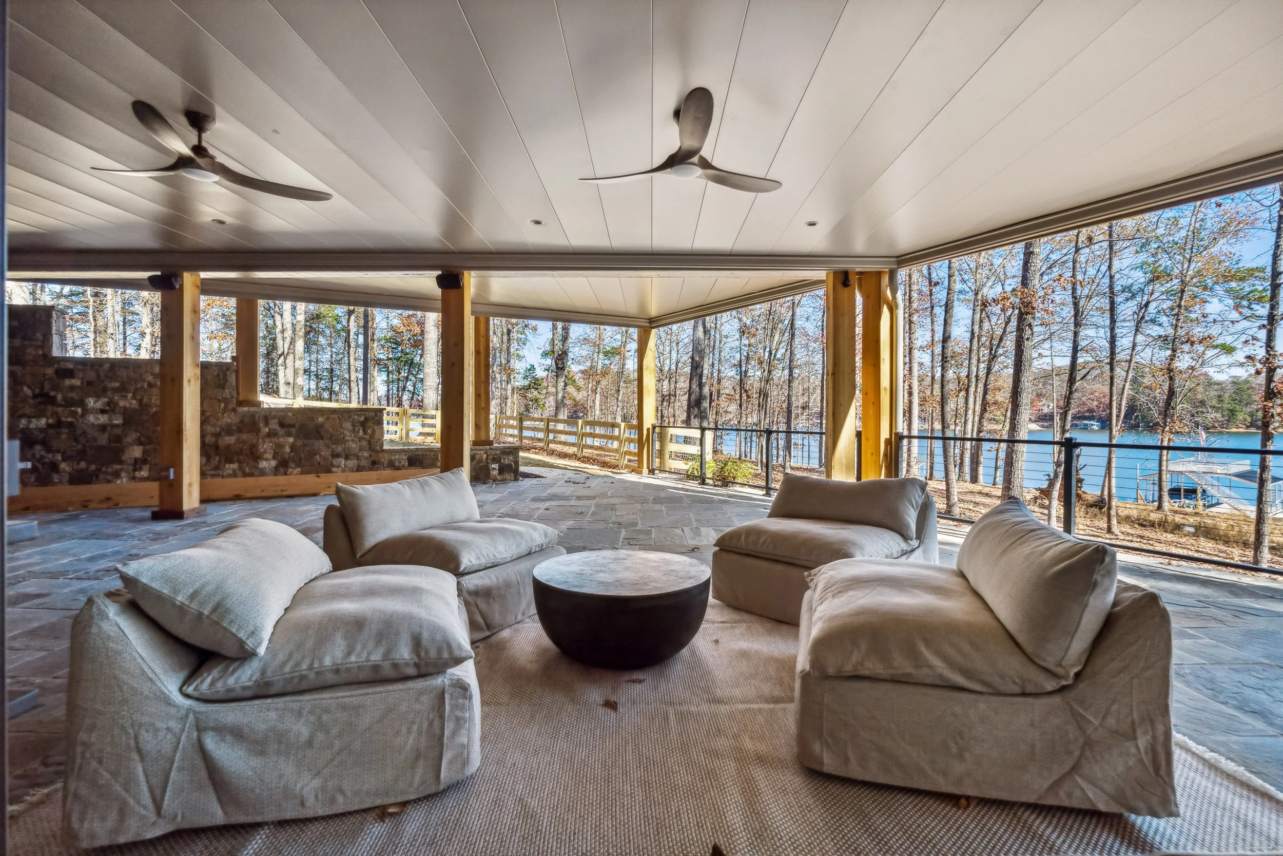 Covered outdoor patio with beige sectional sofa and round black table, stone flooring, wooden posts, and view of lake and trees in fall.