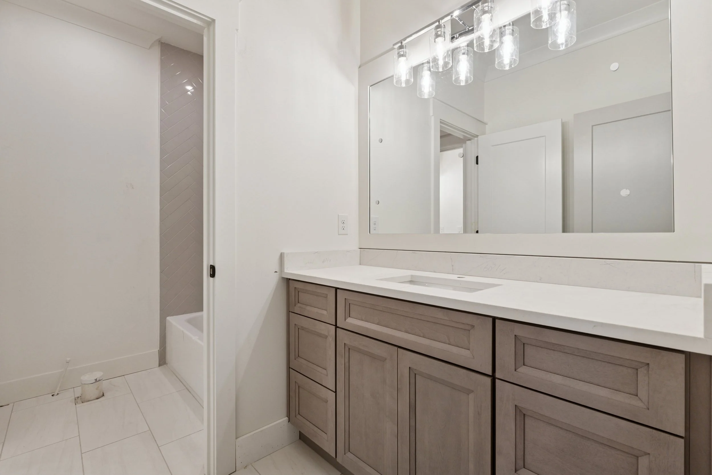 A bathroom vanity with a light-colored wood base, a white marble countertop, and a large mirror. A modern light fixture with multiple transparent glass shades hangs above the mirror. There is a door to the left, leading to a shower area with a white 