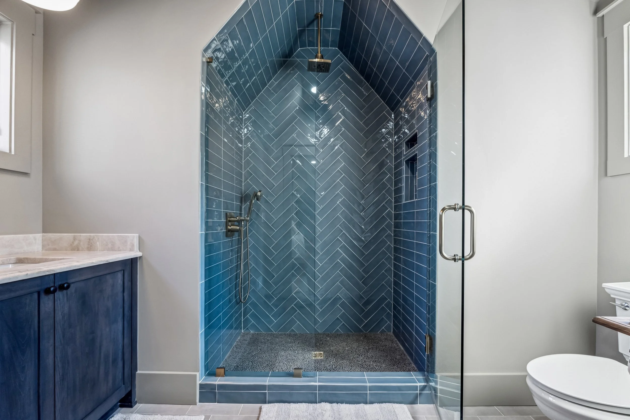Modern bathroom with a walk-in shower enclosed by glass, featuring dark blue subway tiles in a herringbone pattern, a ceiling-mounted shower head, a handheld shower attachment, and a textured floor. To the left is a blue vanity with a marble countert