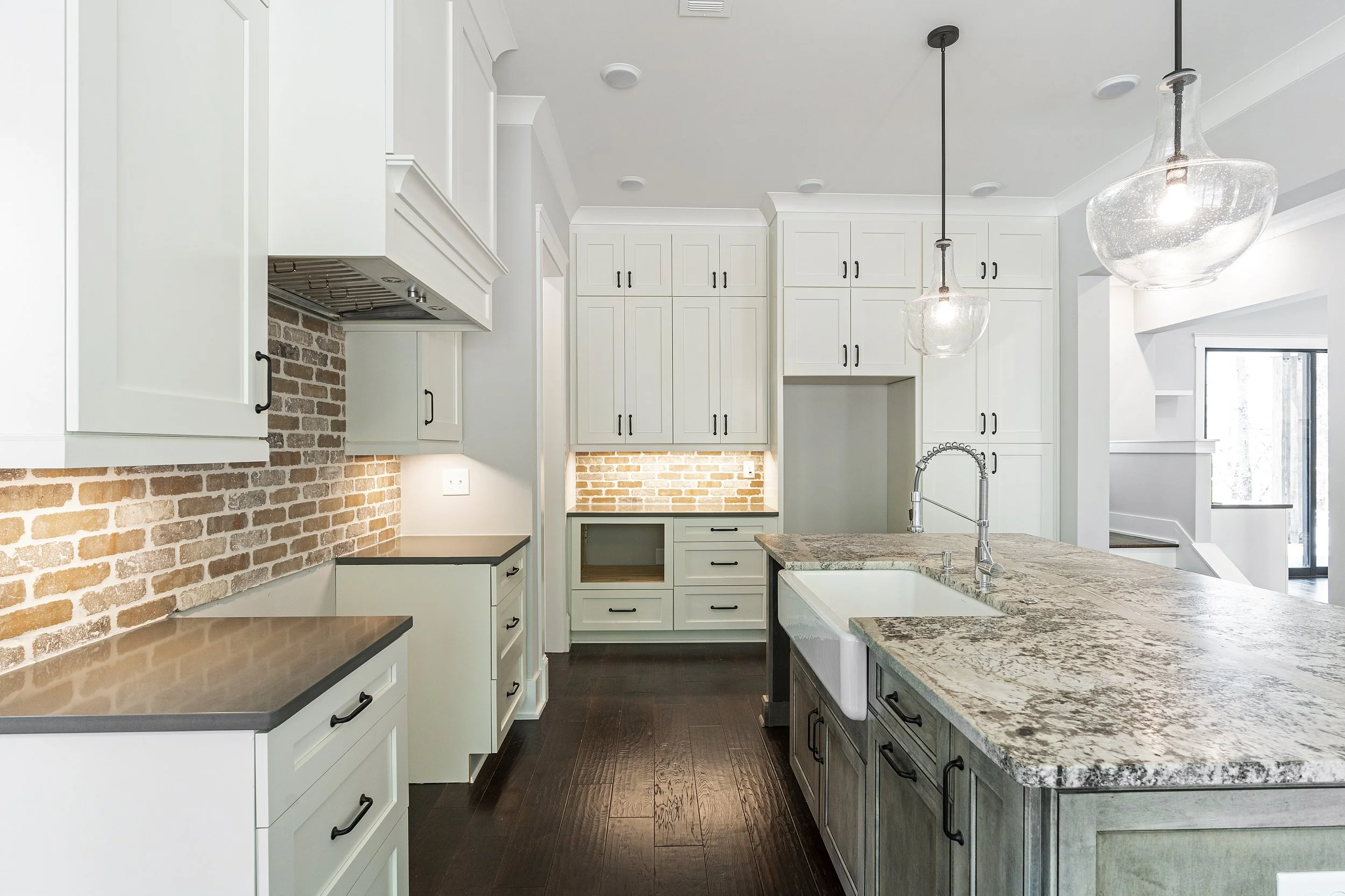 Modern kitchen with white cabinets, brick backsplash, granite countertops, dark wood flooring, and hanging glass pendant lights.