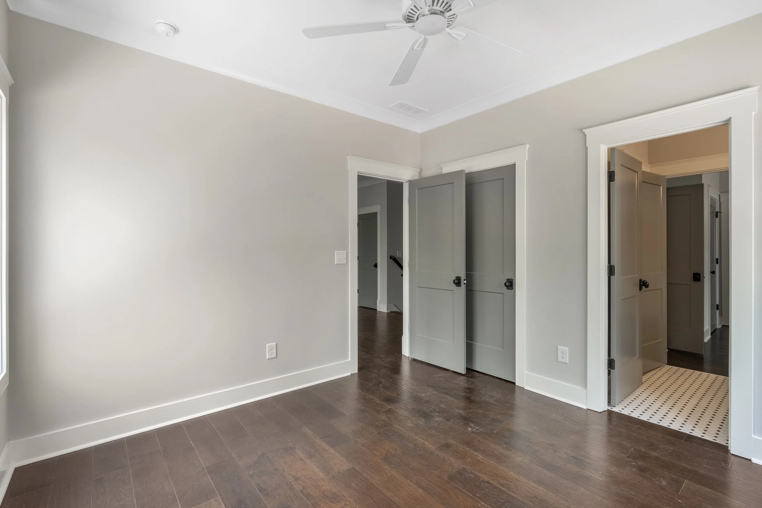 Empty room with white walls, dark wood flooring, and open doors leading to other rooms, with ceiling fan and white trim.