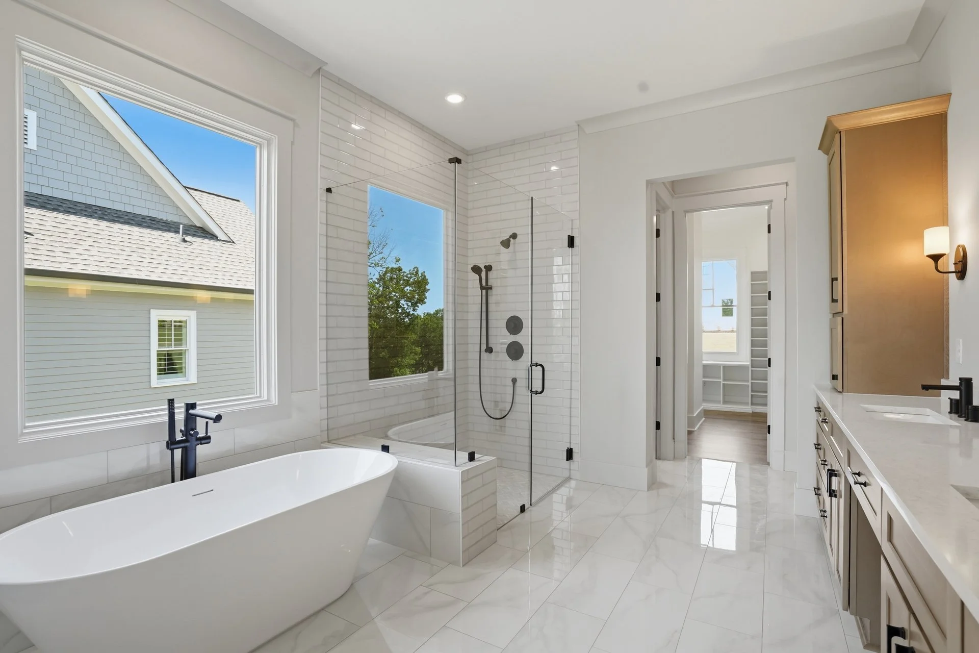 Modern bathroom with white bathtub, walk-in shower with glass enclosure, white brick tile walls, large windows showing neighboring house, wooden cabinet, and black fixtures.