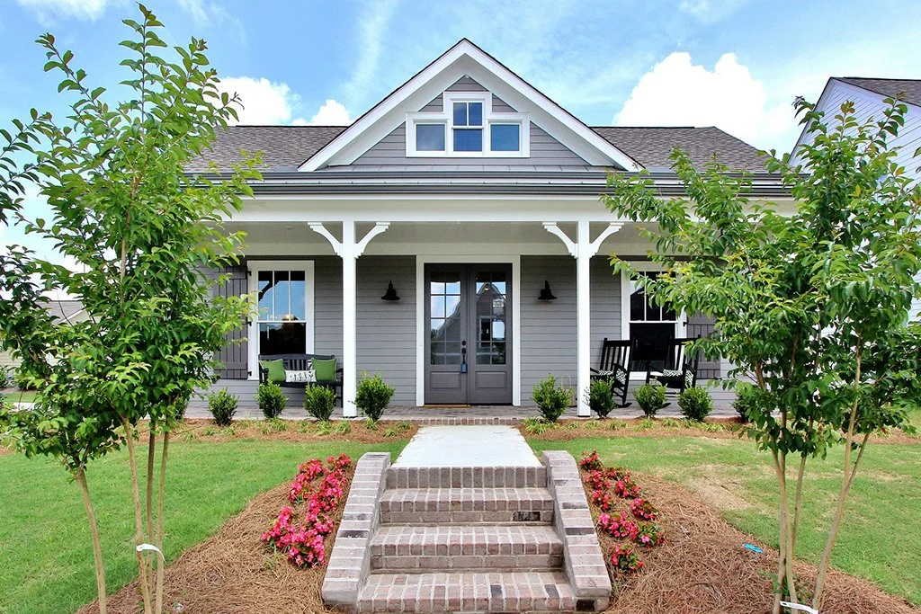 Front view of a modern gray house with a porch, stairs, and landscaping including trees and flowers.