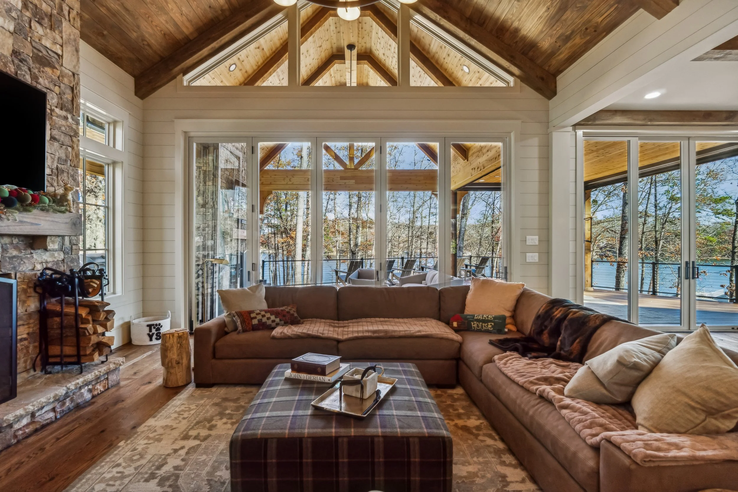 Living room with a large sectional sofa, plaid ottoman, wood flooring, stone fireplace, and large windows with a view of trees outside.