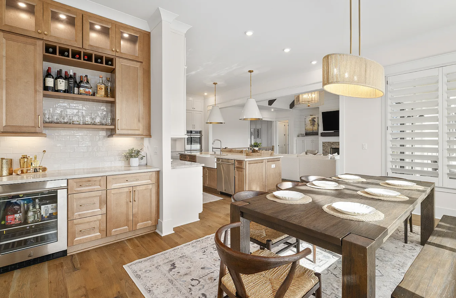 Open-concept kitchen and dining area with wooden cabinetry, a large dining table set for six, and a view into living room with a fireplace and television. Modern lighting and white walls add brightness.