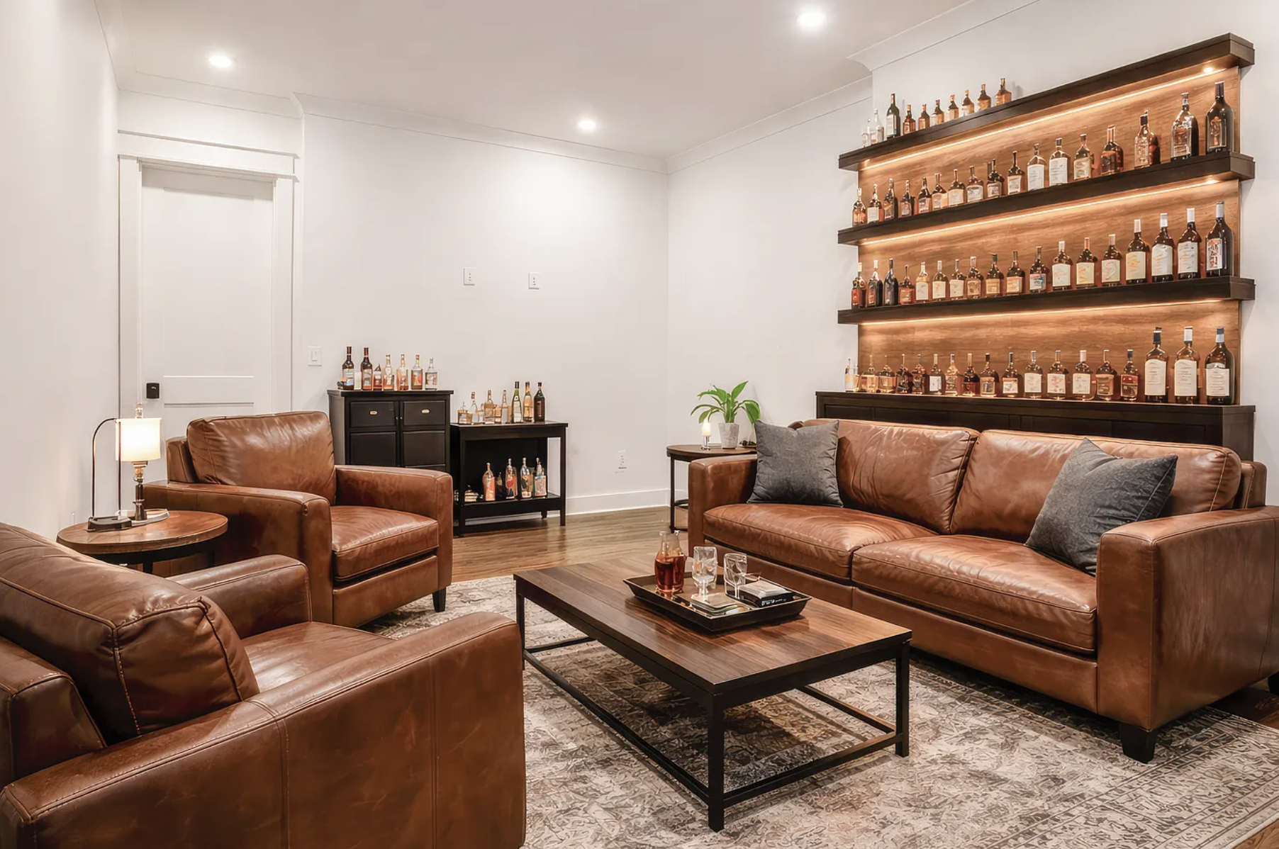 Living room with leather sofas, wooden coffee table, and a wall-mounted liquor shelf with bottles. There is a small black cabinet with bottles and a larger black sideboard in the corner. A lamp and a potted plant are on side tables, and the room has 