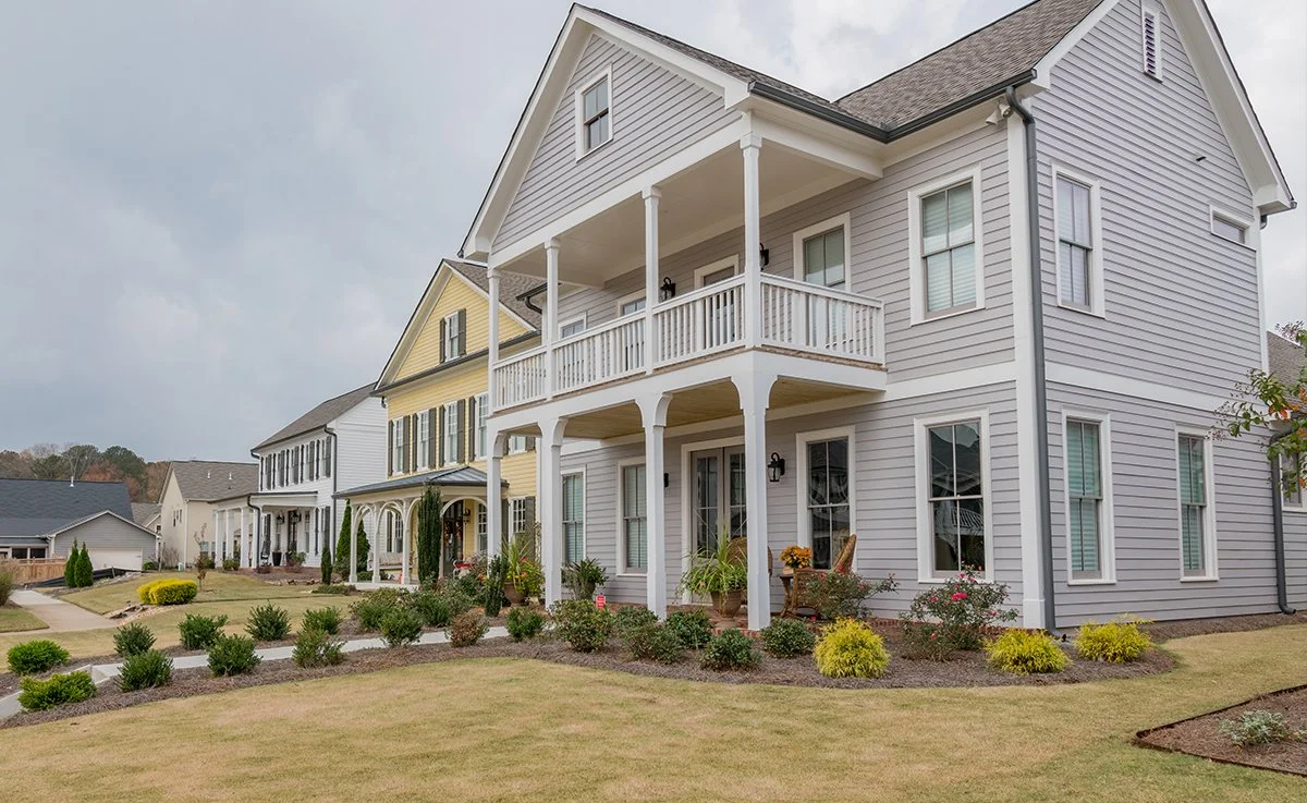 Multiple modern houses with well-maintained lawns and landscaping, featuring a gray house with a front porch and balcony, set in a suburban neighborhood.
