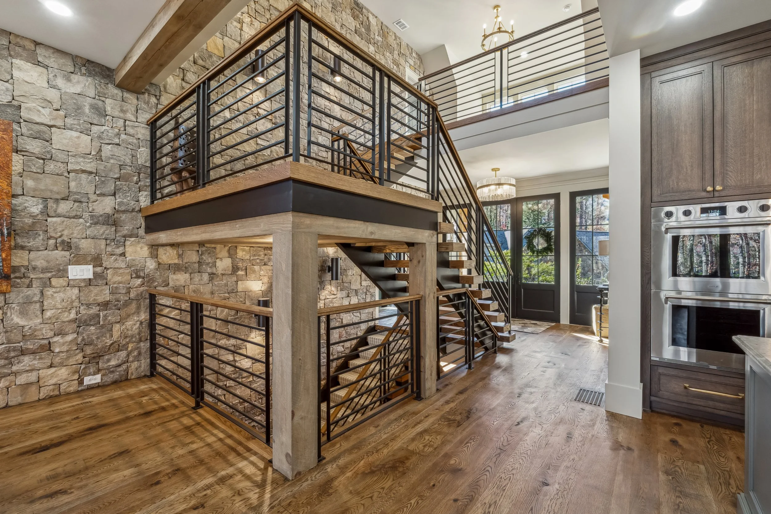 Interior view of a house with a spiral staircase, stone wall, wooden floors, and a kitchen area with cabinetry and appliances.