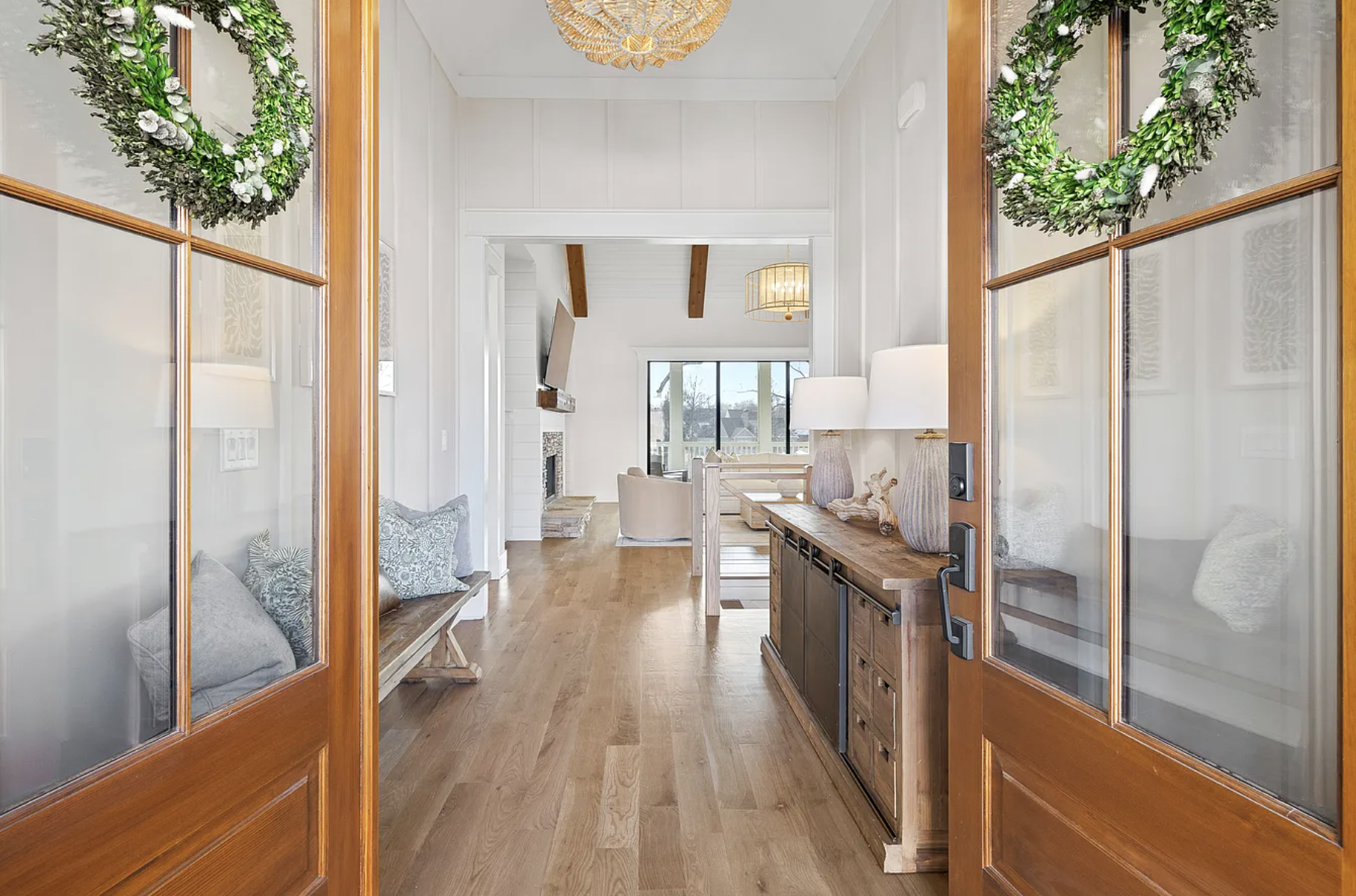 View through wooden-framed glass double doors into a bright, modern living room with a high ceiling, large window, fireplace, and hardwood floor, decorated with wicker and natural decor.