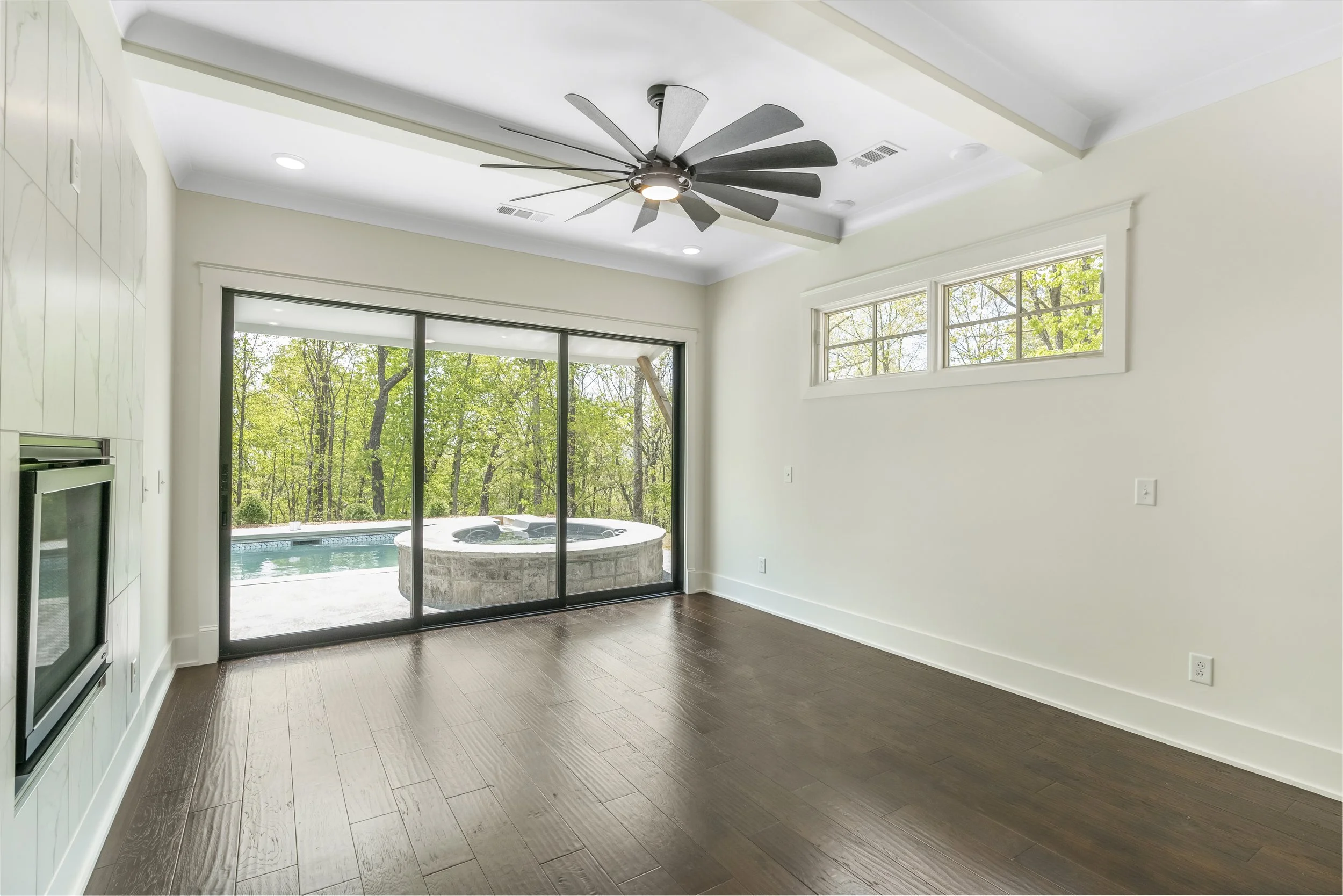 Inside a room with dark wood flooring, white walls, and a ceiling fan, next to a large glass sliding door that opens to an outdoor hot tub and view of trees.