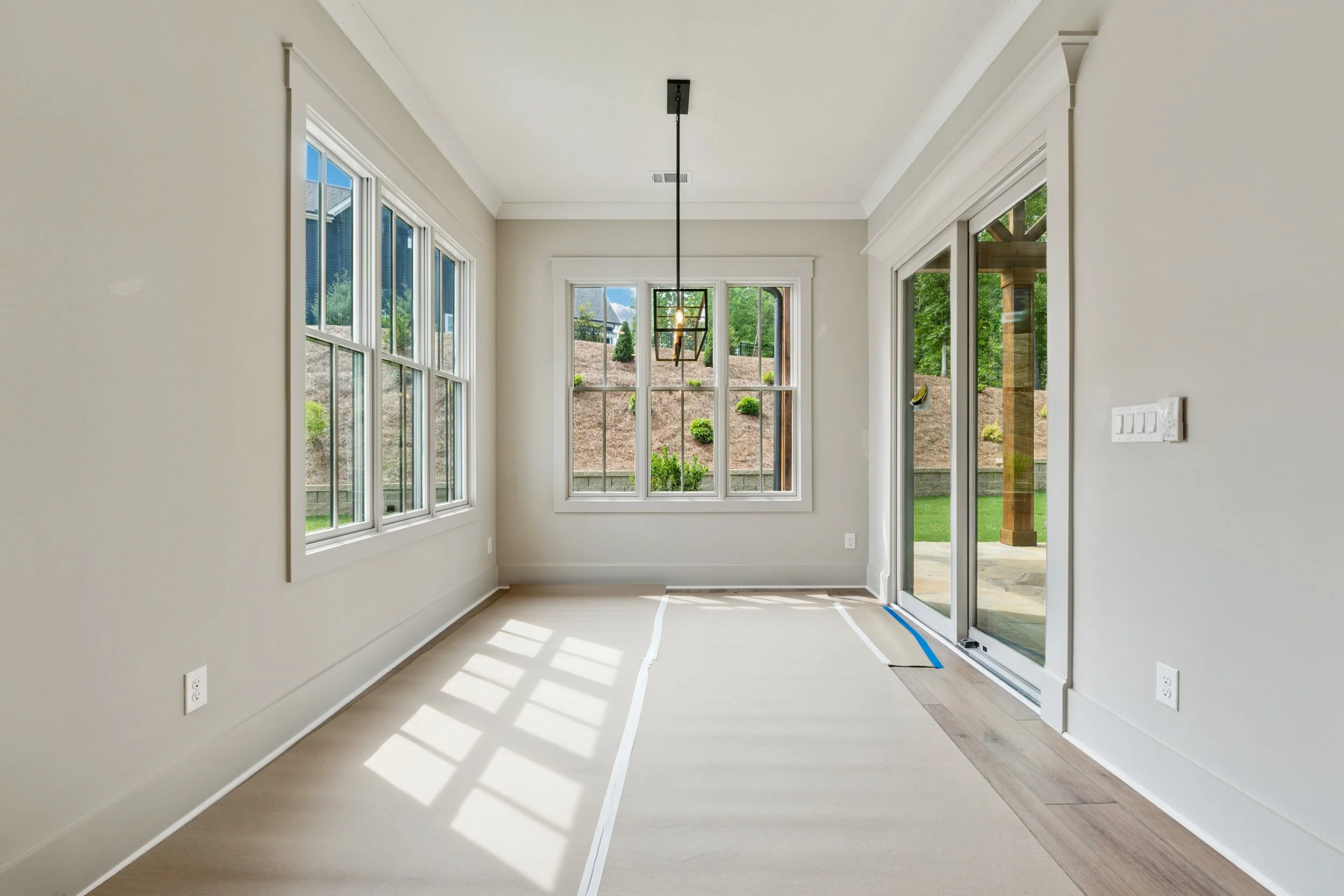 Empty room with large windows and sliding glass door, sunlight creating shadows on light-colored floor.