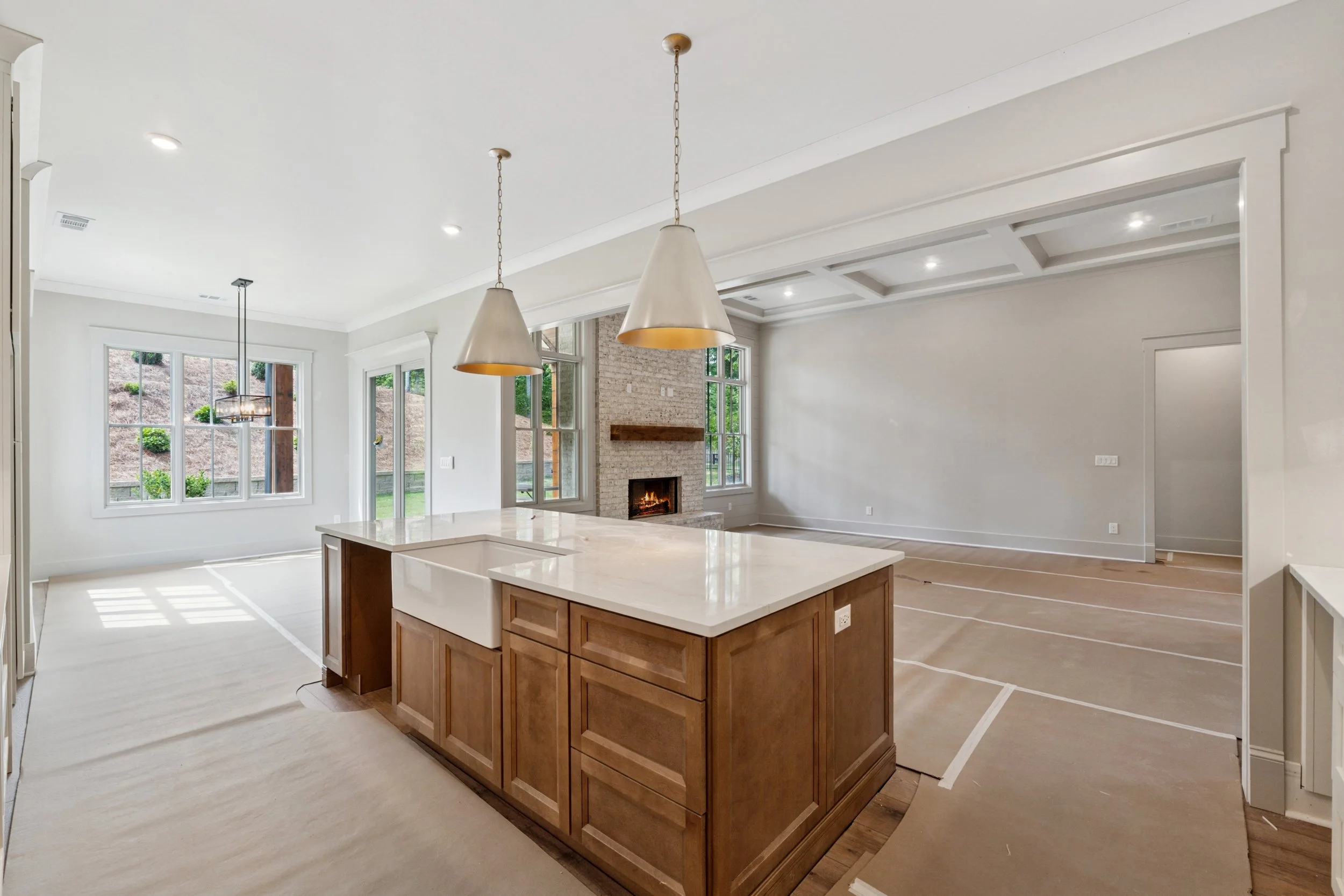 Open-concept kitchen and living room with large windows, hardwood flooring, a brick fireplace, modern pendant lights, and an island with a white countertop.