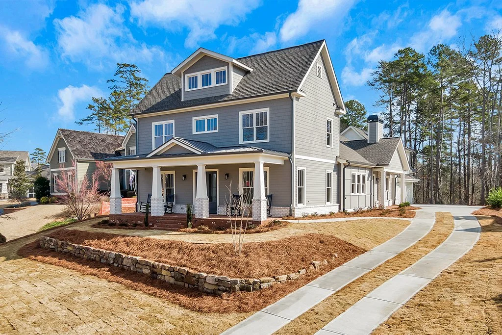 A modern, gray two-story house with a front porch, surrounded by a freshly landscaped yard with a stone-lined flower bed and a curved concrete driveway under a blue sky.