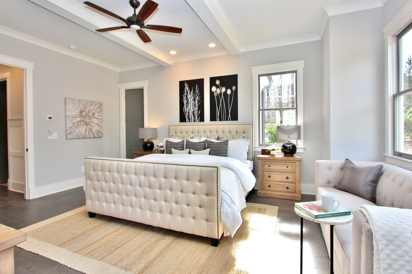 A bedroom with a bed, nightstands, lamps, and wall art, with a window and a ceiling fan.