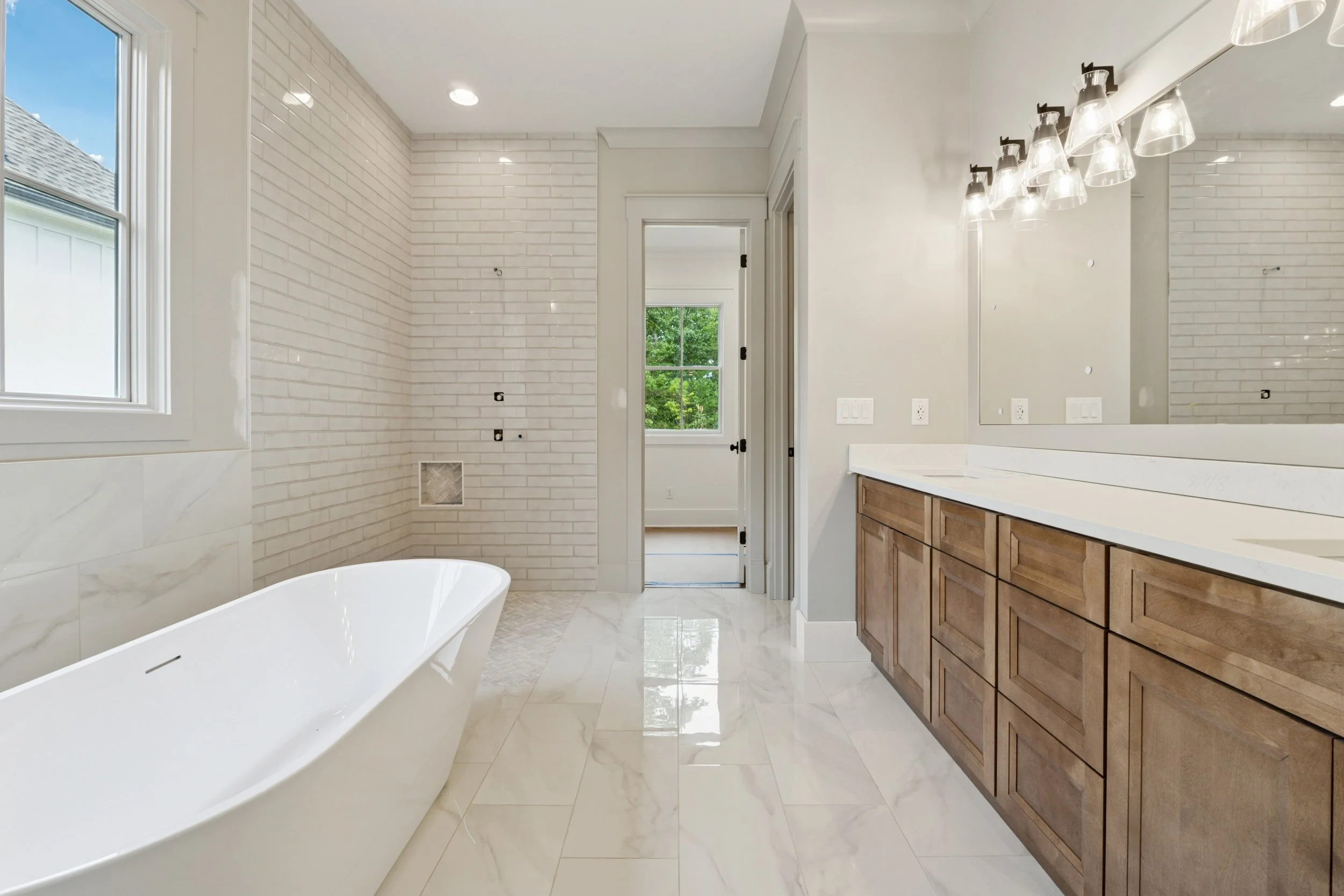 Modern bathroom with white subway tiled walls, marble flooring, a white bathtub, large mirror, wooden vanity, and adjustable lighting fixtures.