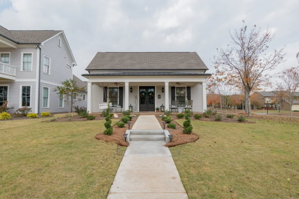 Front view of a house with a white porch, landscaped lawn, small bushes, young trees, and a concrete walkway leading to the entrance. Neighboring houses and fall-colored trees in the background.