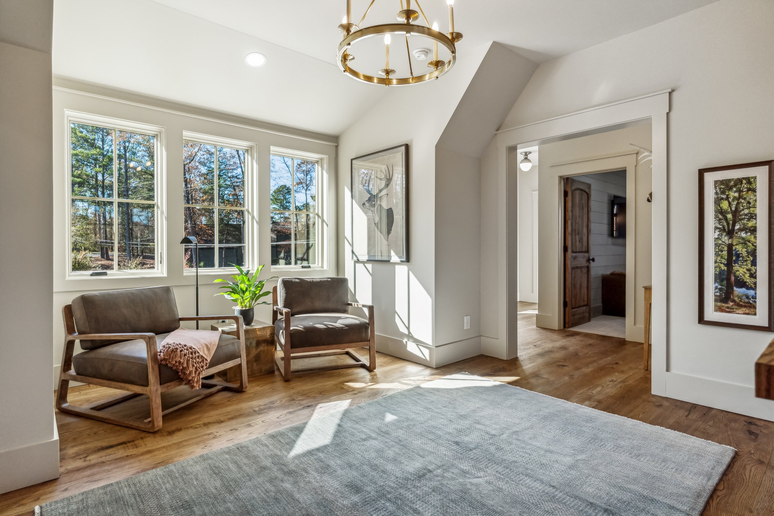 Living room with three large windows, a potted plant, two wooden armchairs with beige cushions, a gray area rug, and a modern gold chandelier hanging from the white ceiling. There is a framed picture of a deer on the wall and an open doorway leading 
