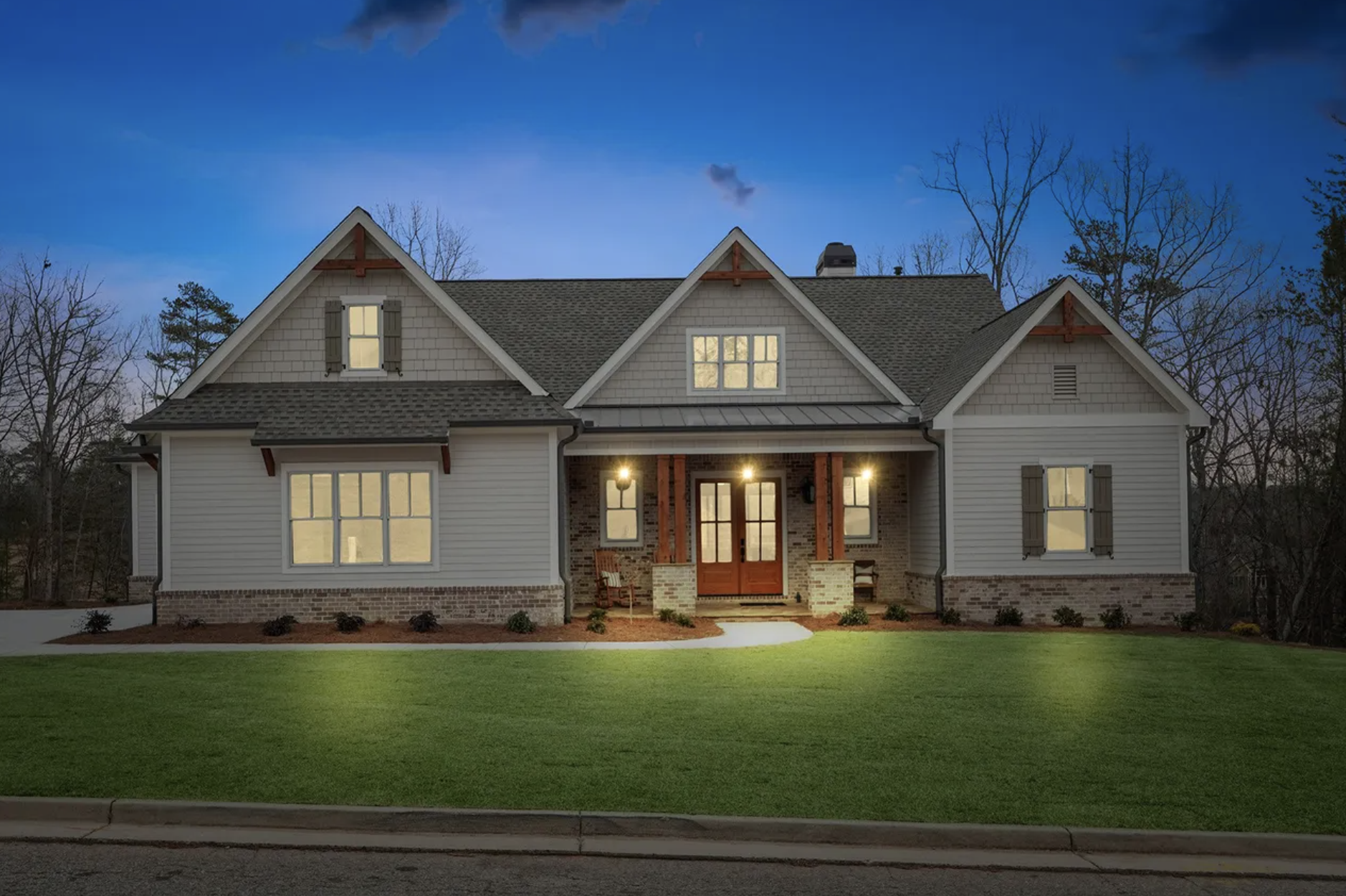 A two-story house with a brick and wood exterior, lit from within, with a well-manicured lawn and a sidewalk in front. The house has dormer windows, a covered porch with two chairs, and a dark shingled roof. The sky is dusk or early evening.
