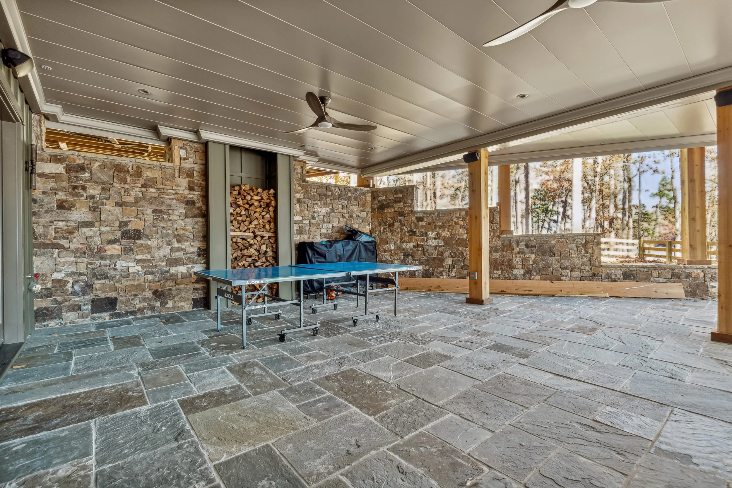 A covered outdoor patio with stone tile flooring, a stone wall with firewood storage, a ceiling fan, and large open windows showing a wooded landscape.