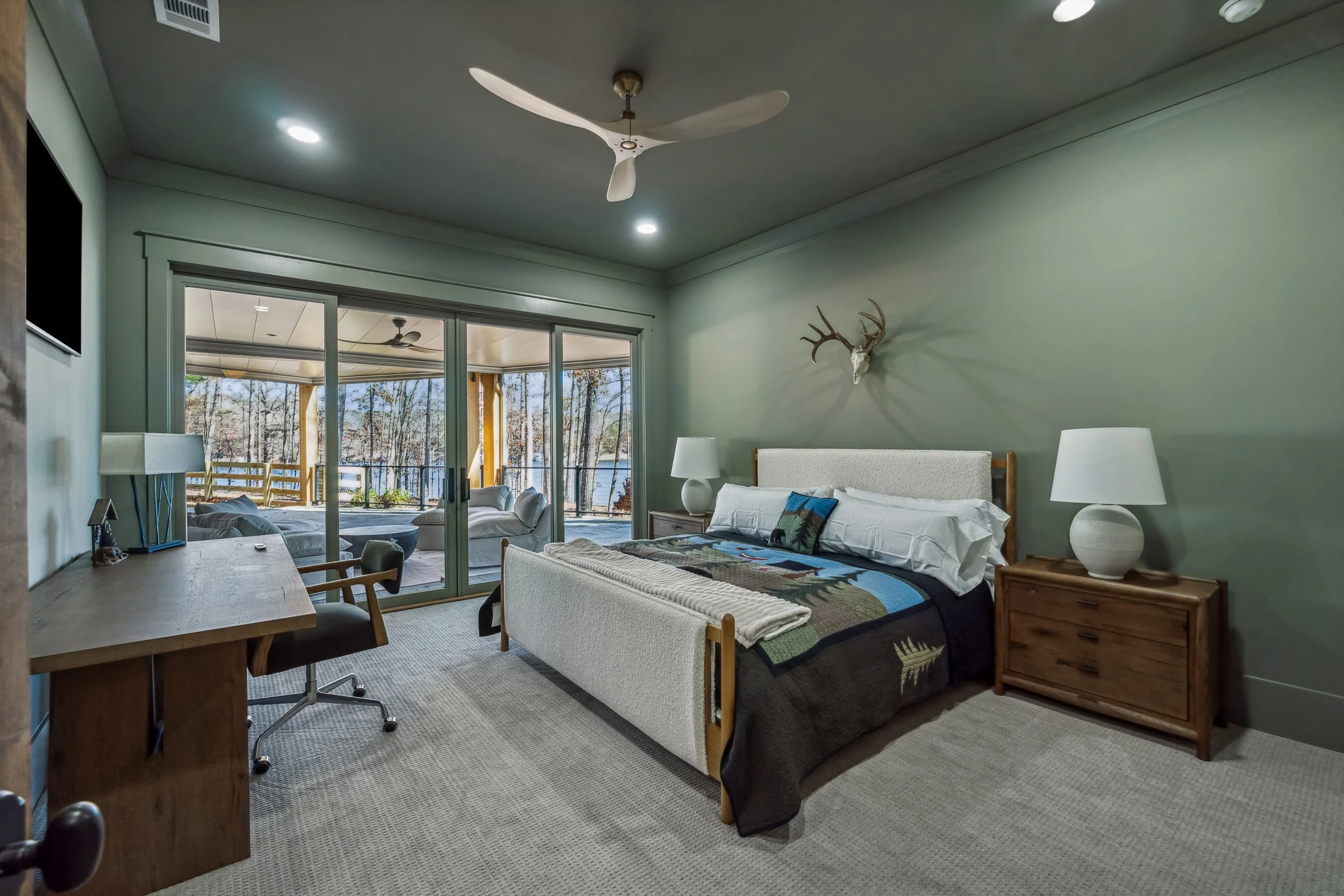 A bedroom with a large bed, two white nightstands with lamps, a deer skull with antlers mounted on the green wall, and sliding glass doors leading to a patio with outdoor seating and trees.