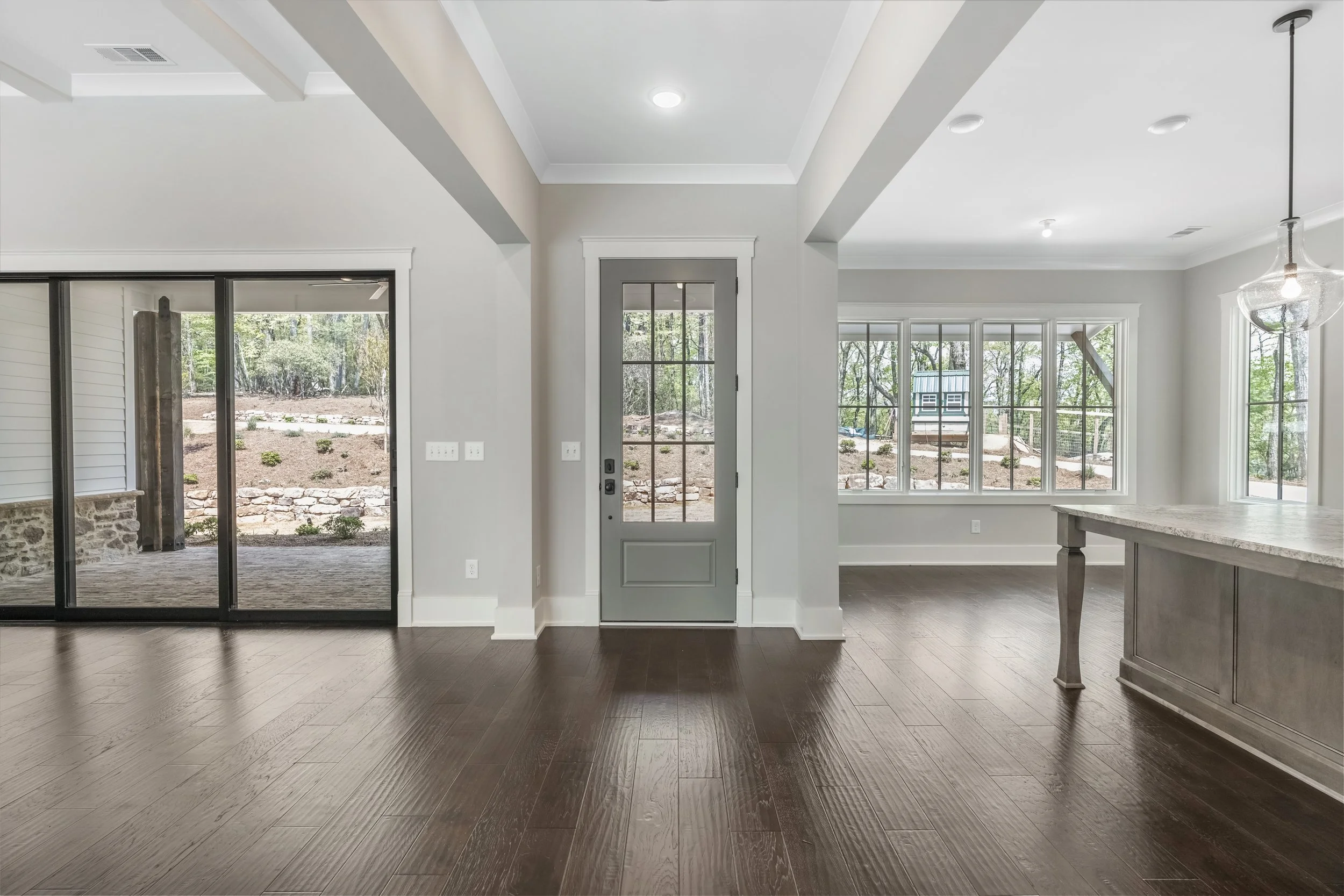 Interior view of a modern house with white walls, large windows, and a wooden floor. There is a sliding glass door leading outside, and a ceiling light and hanging pendant light are visible.