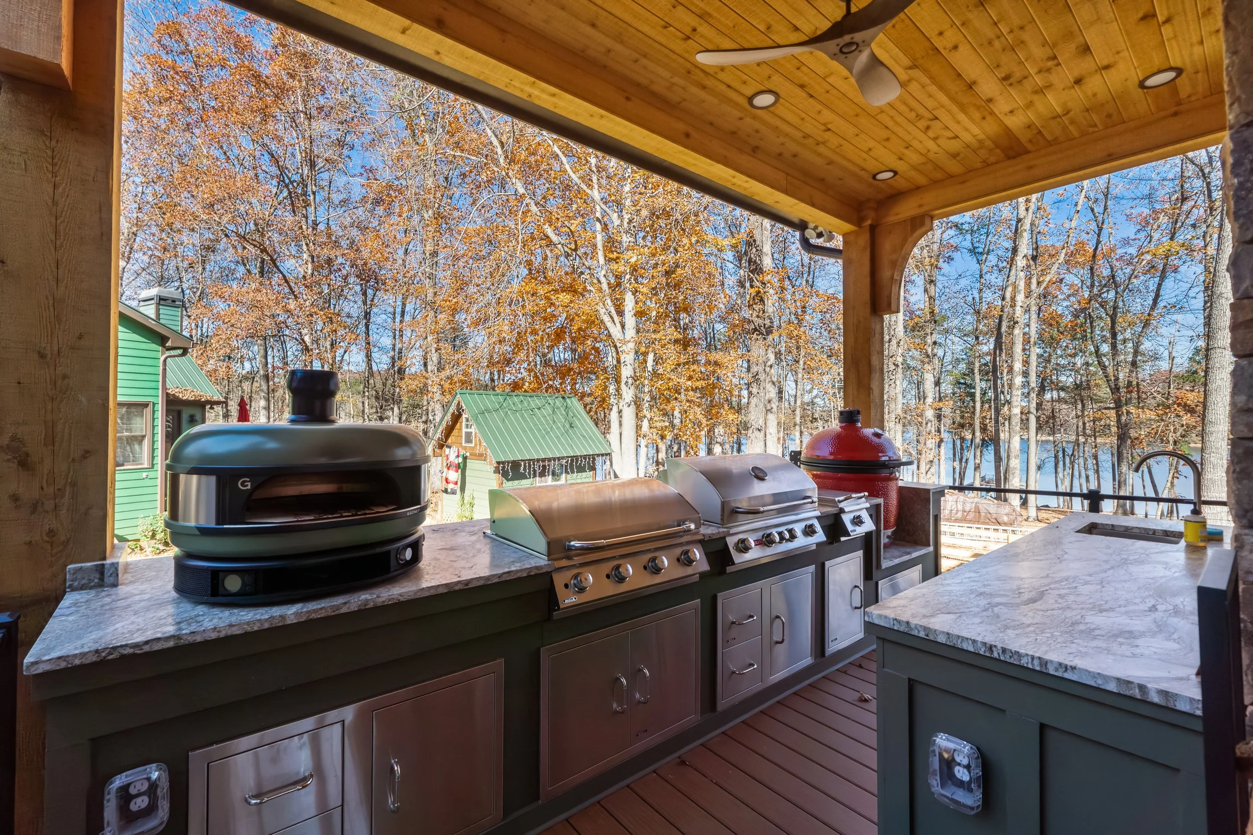 Outdoor kitchen on a wooden deck with grill and smoker, surrounded by trees with autumn leaves, a green house in the background, and a clear blue sky.