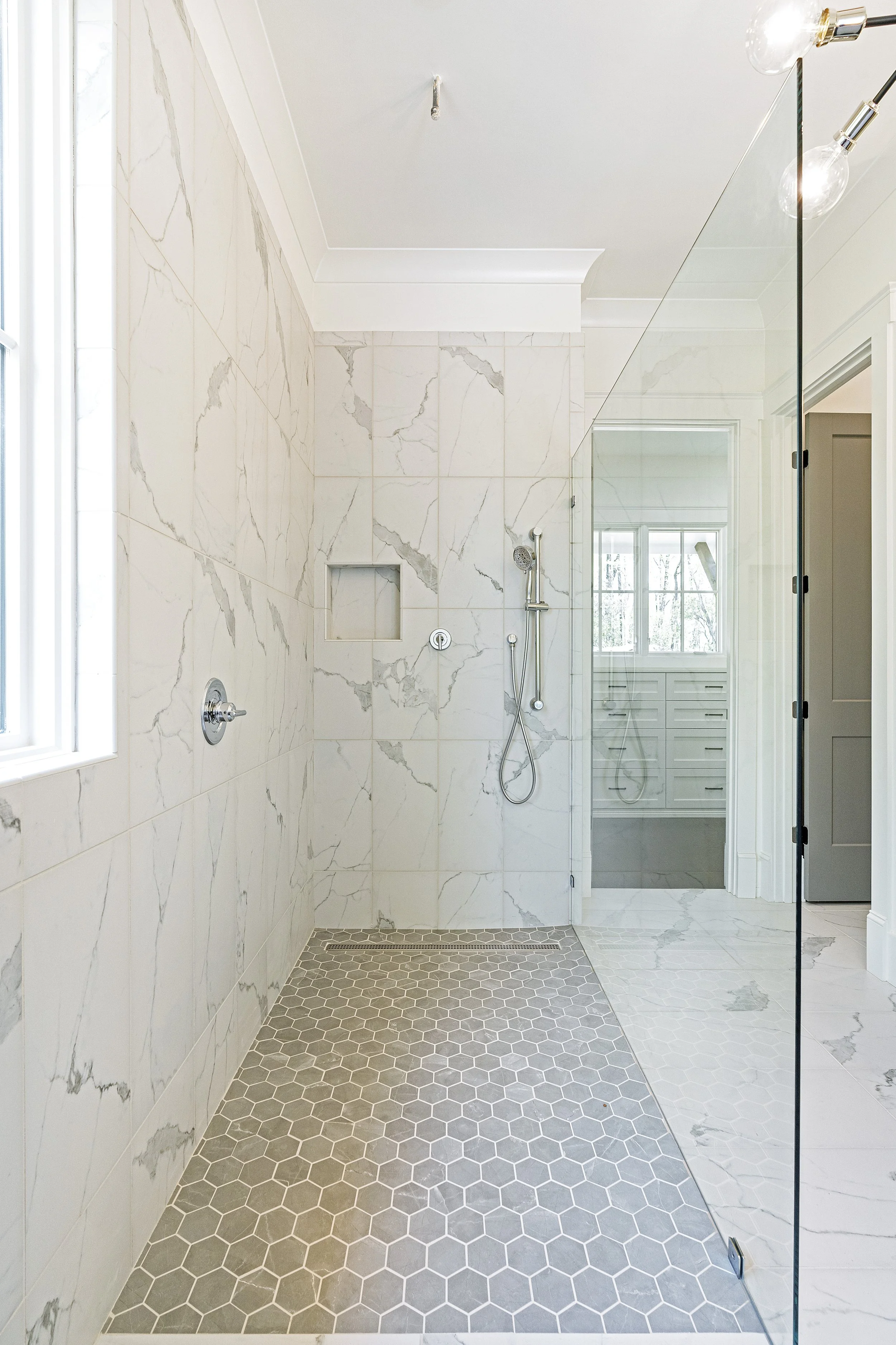 Modern shower area with marble wall tiles, a glass partition, a rain shower head, and a window letting in natural light.