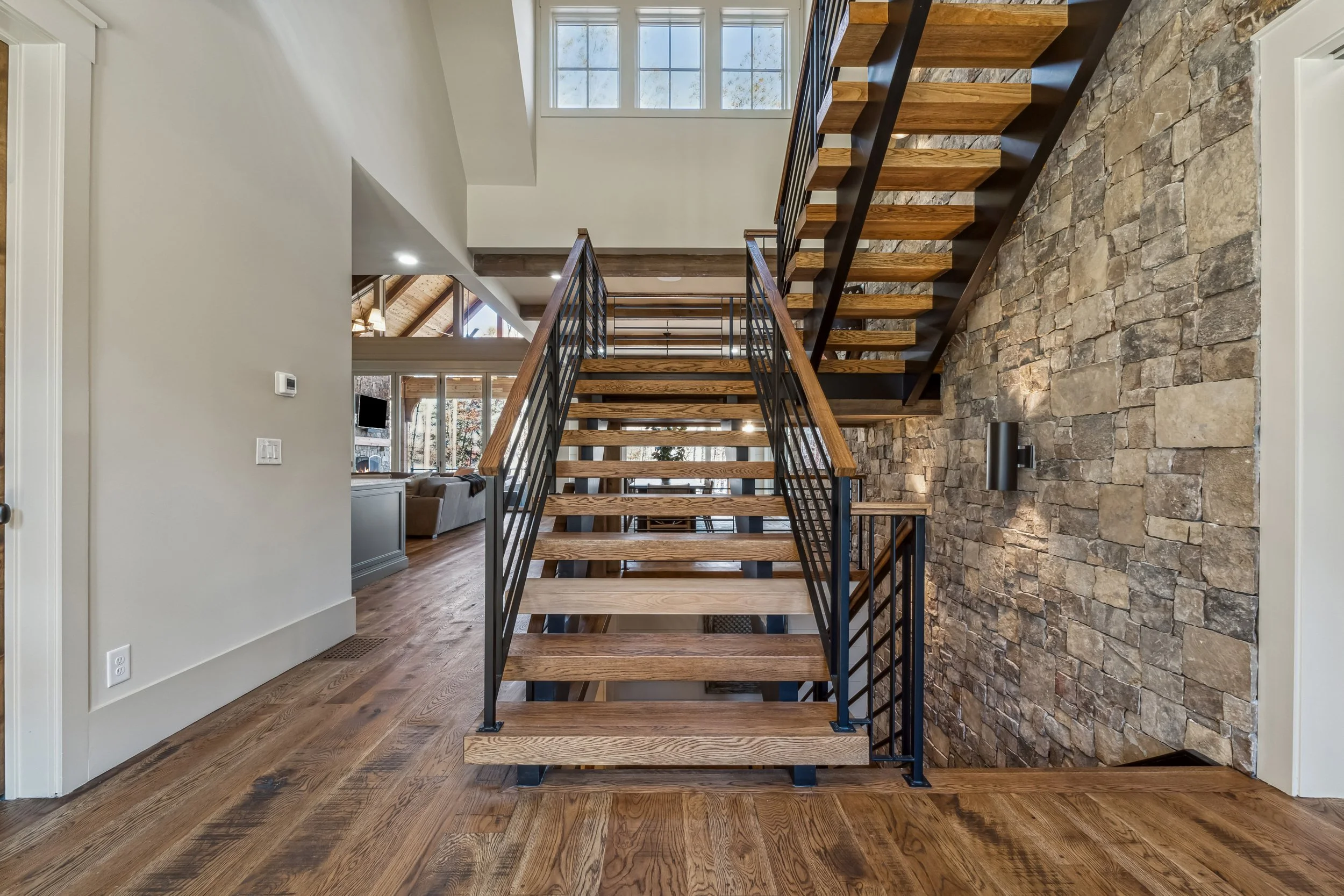 Interior view of a house with a staircase featuring wooden steps and iron railings, a stone accent wall, hardwood flooring, large windows letting in natural light, and a partial view of a living area with a couch and a TV.