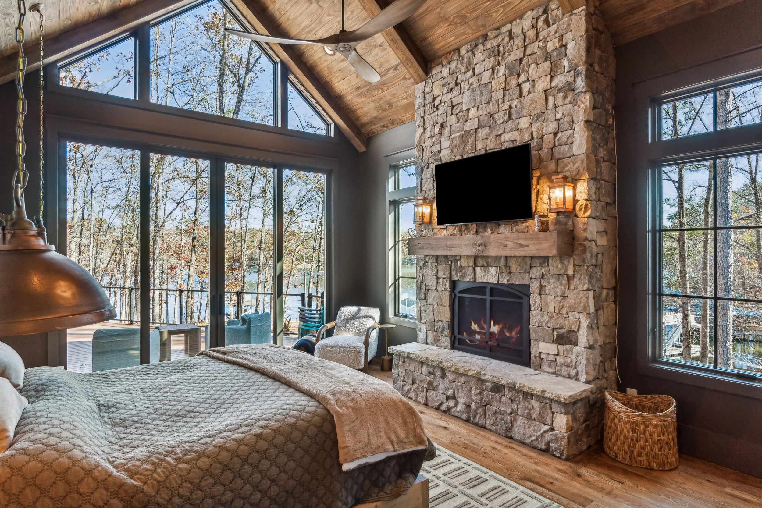 Living room with large windows showing a view of trees and water outside, stone fireplace with a fire, wall-mounted TV, wooden ceiling with a ceiling fan, armchair, bed in the foreground, and a wicker basket.